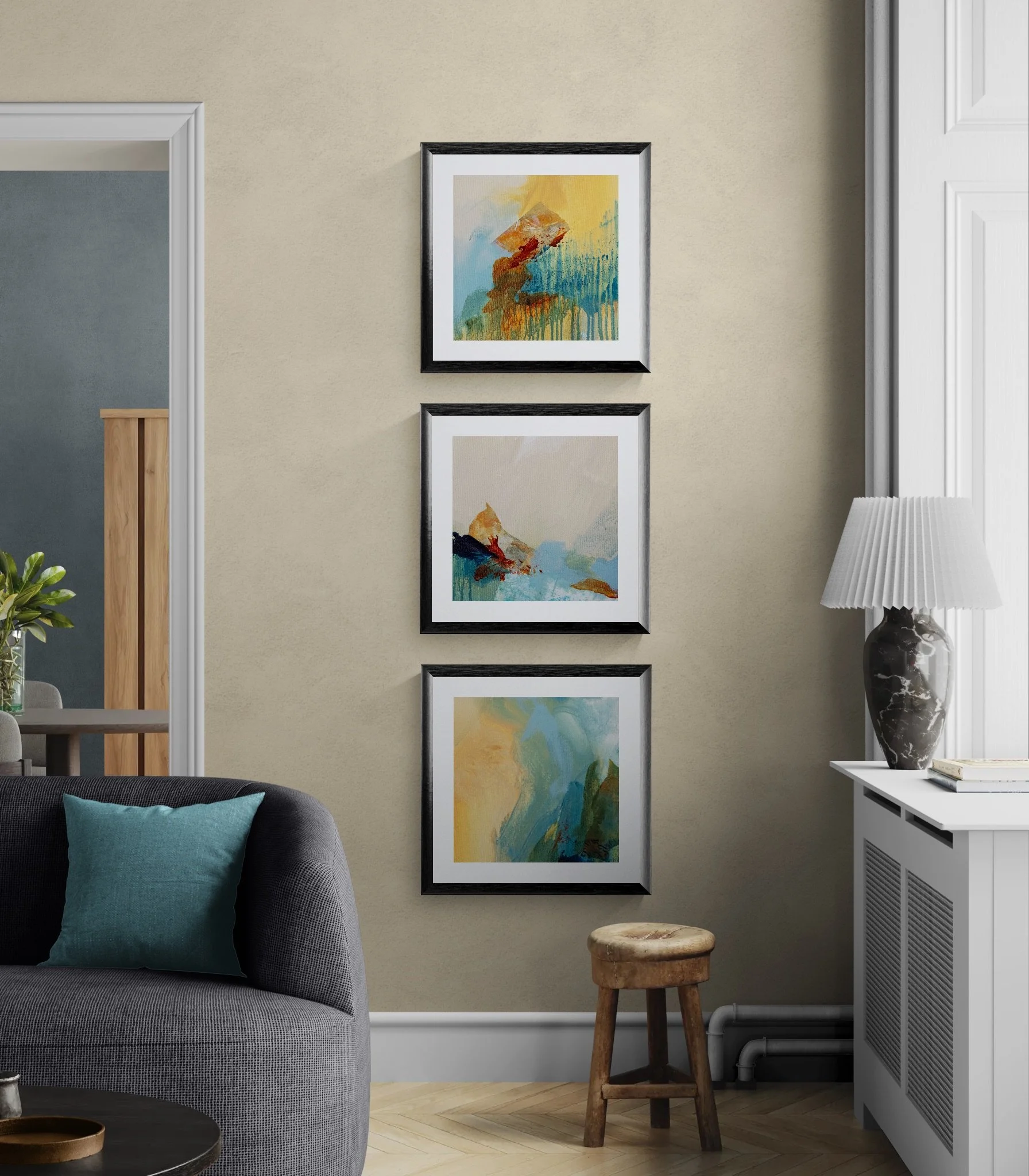 Three abstract paintings hanging vertically on a beige wall in a living room. Part of a gray sofa with a turquoise pillow is visible on the left. On the right, there is a white sideboard with a large black and white marble vase lamp and a stack of books. A small wooden stool is in front of the sideboard.