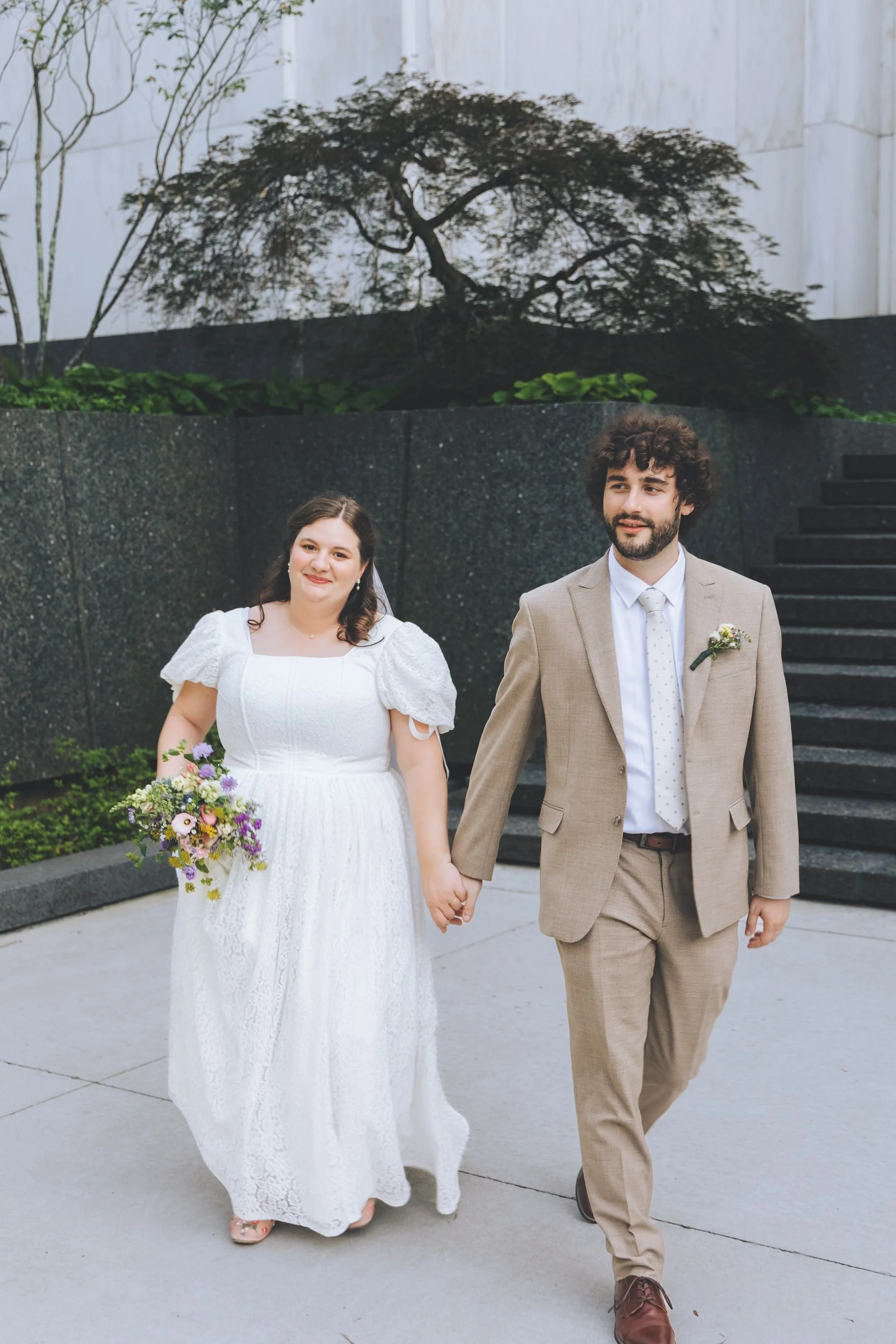 A bride and groom walking hand in hand outdoors, with the bride in a white dress holding a bouquet and the groom in a beige suit, near stairs and a wall with a decorative tree.