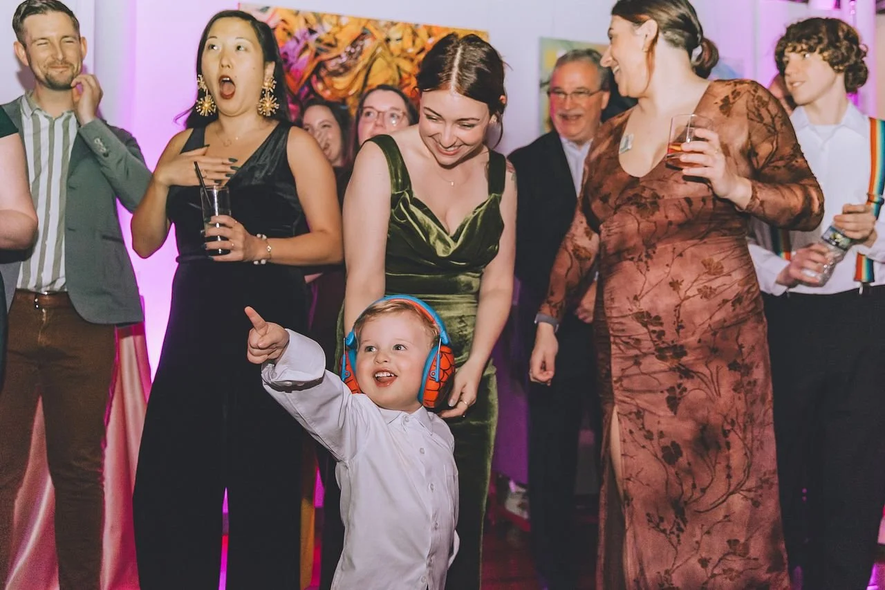 A group of adults and a child at a lively indoor celebration, with some holding drinks and one child wearing headphones, smiling and pointing.