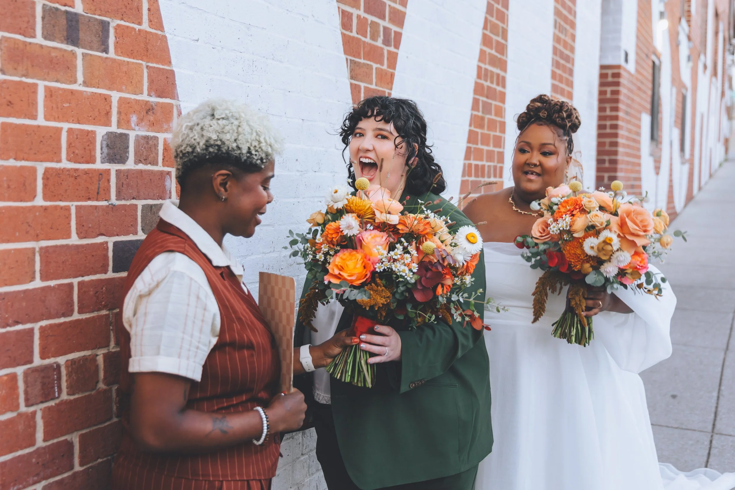Two women in wedding dresses holding bouquets of flowers and smiling with a woman in a brown vest and striped shirt outside against a brick wall.