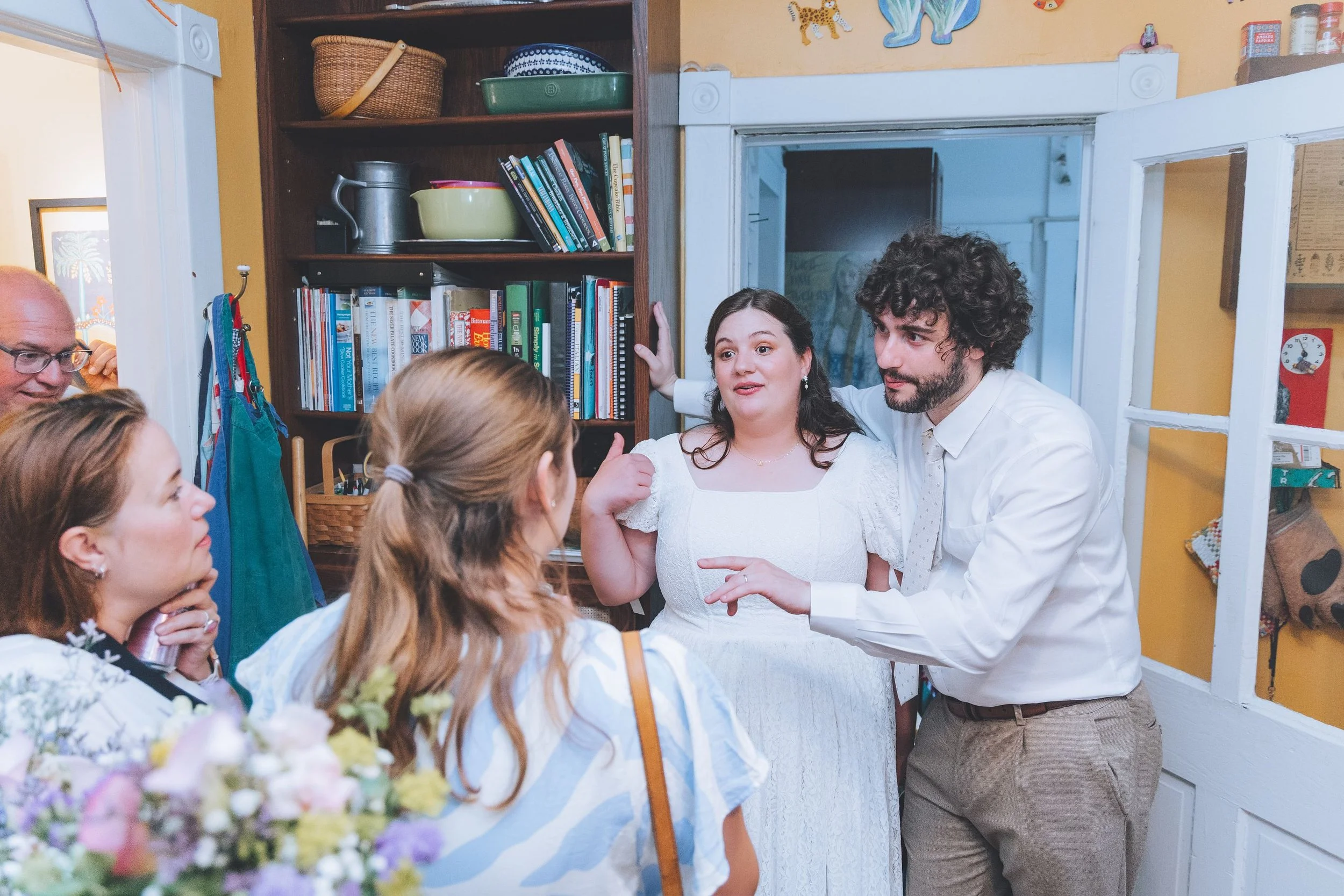 Group of five people having conversation in a cozy room, with bookshelves, a yellow wall, and a wall clock in the background.