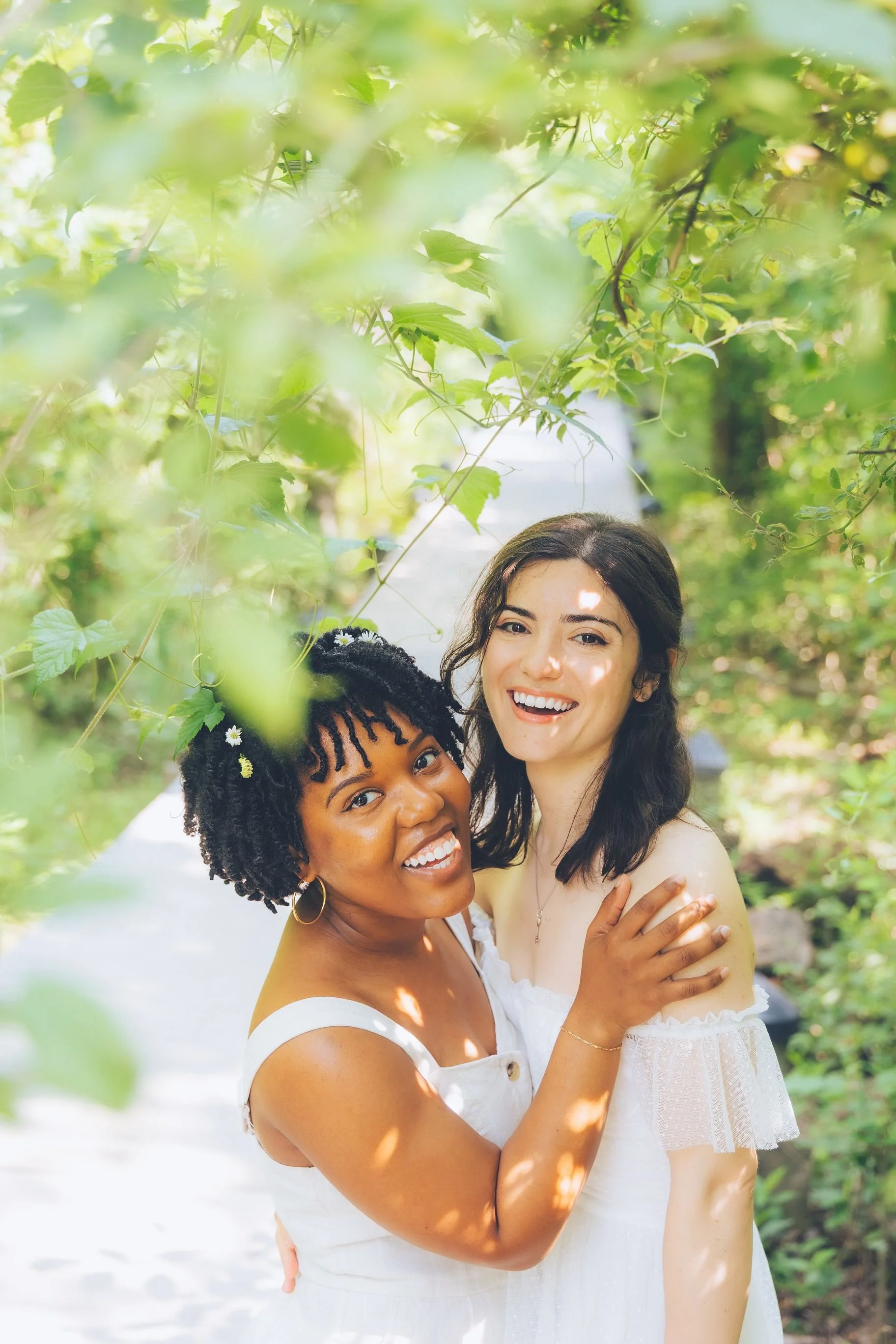 Two women smiling and embracing outdoors among green foliage, with sunlight filtering through leaves.