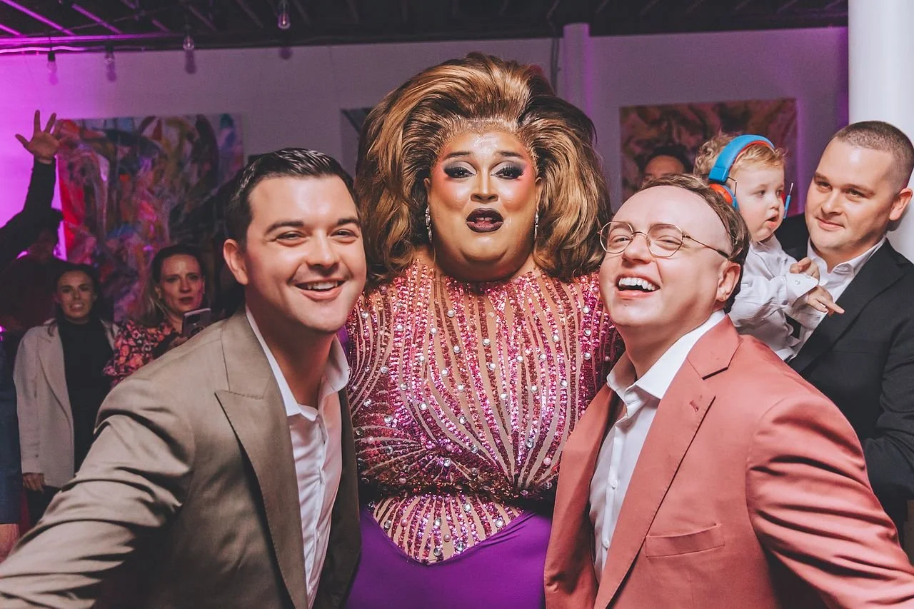 Group of people at a lively party, including three men in suits, a performer in a sparkly pink dress with voluminous hair, and a young girl with headphones, with colorful lighting and artwork in the background.
