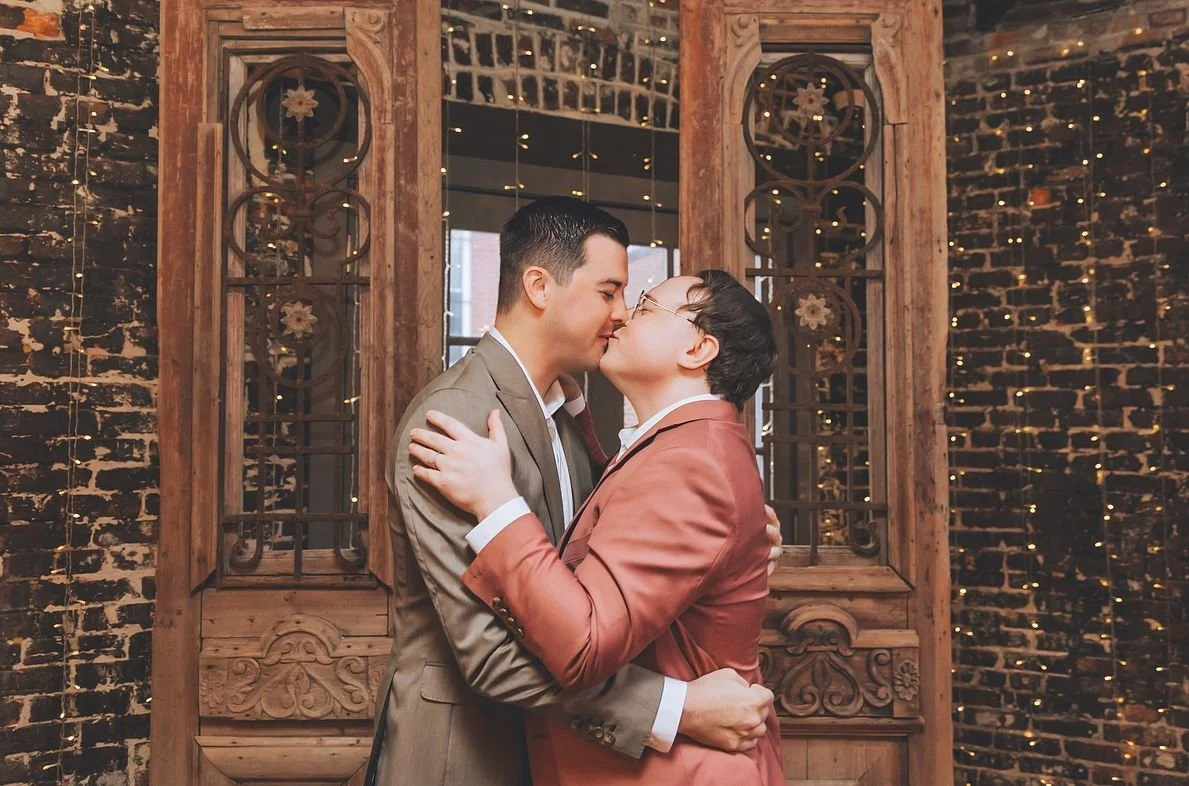 Two men in suits embracing and kissing in front of a decorative wooden wall with string lights.