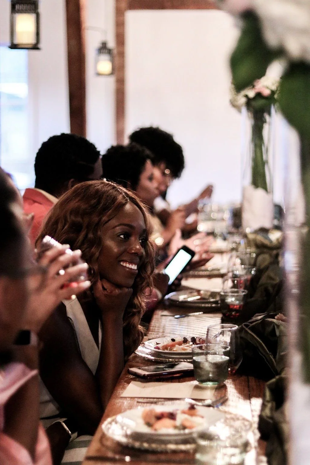 A group of people sitting at a long dinner table, some are looking at their phones and smiling.