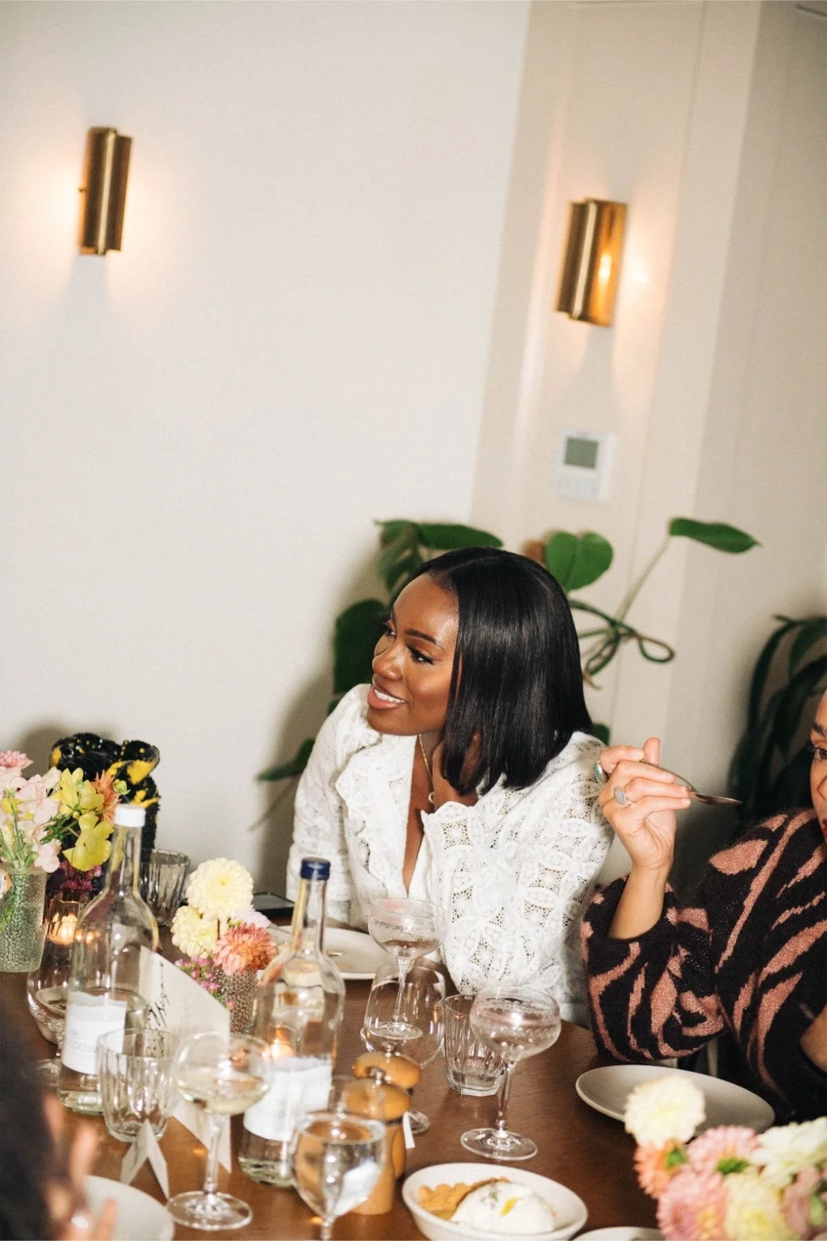A woman smiling and sitting at a dinner table with flowers and drinks, wearing a white lace top.