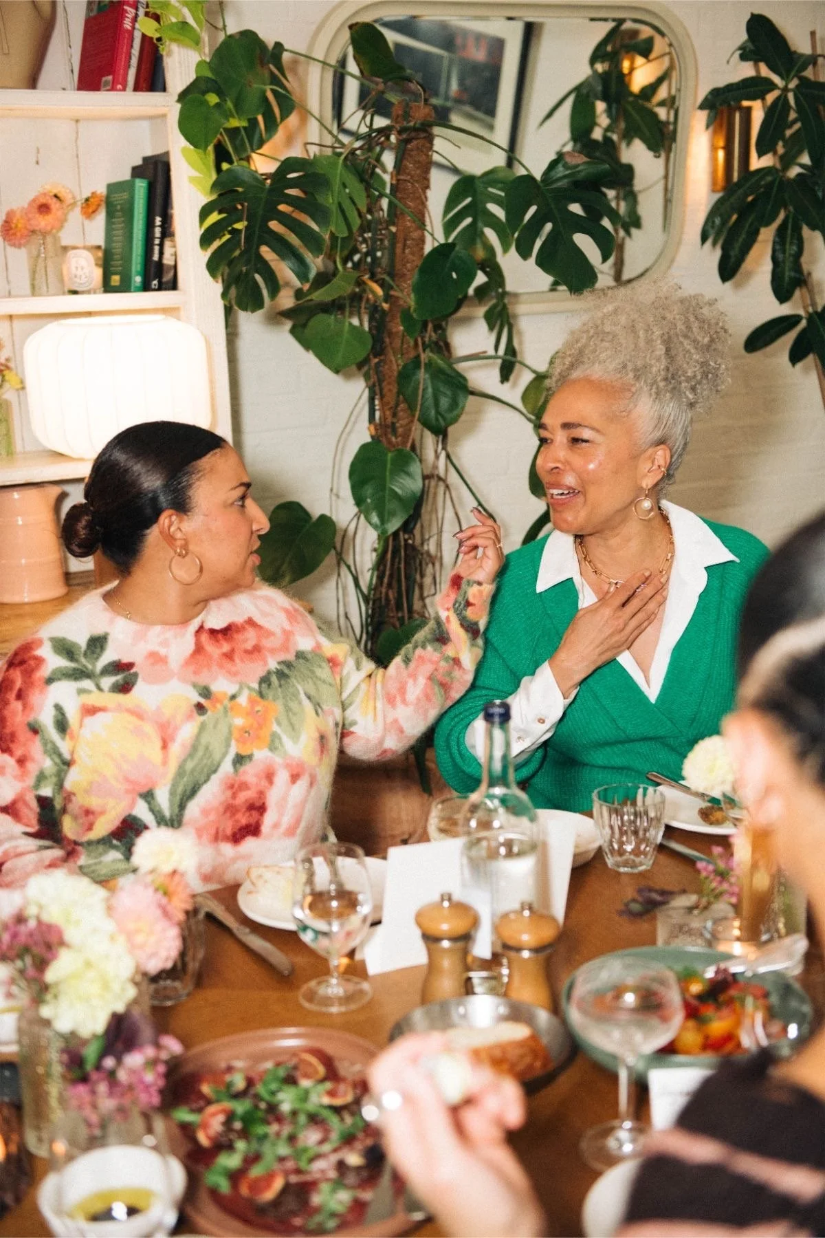 Two women talking at a dinner table decorated with flowers. The woman on the left wears a colorful floral sweater, and the woman on the right, with gray hair, wears a green sweater. Behind them is a leafy plant and a mirror.