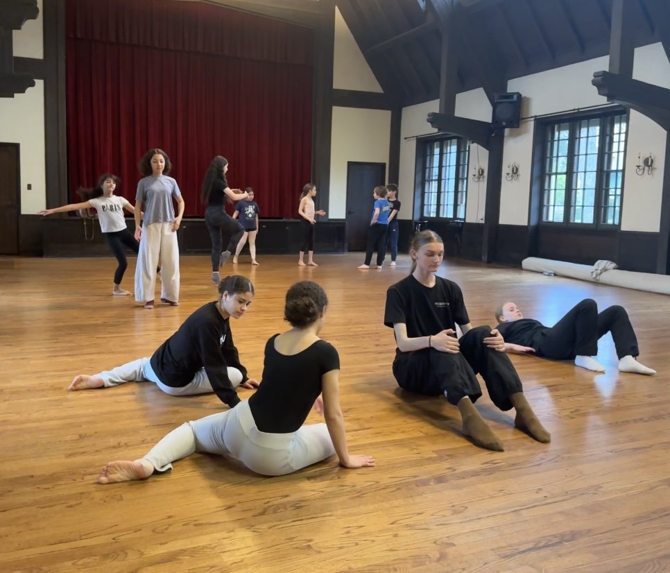 Young ballet dancers practicing stretches and poses in a spacious dance studio with wooden floors, large windows, and a red curtain stage at the back.