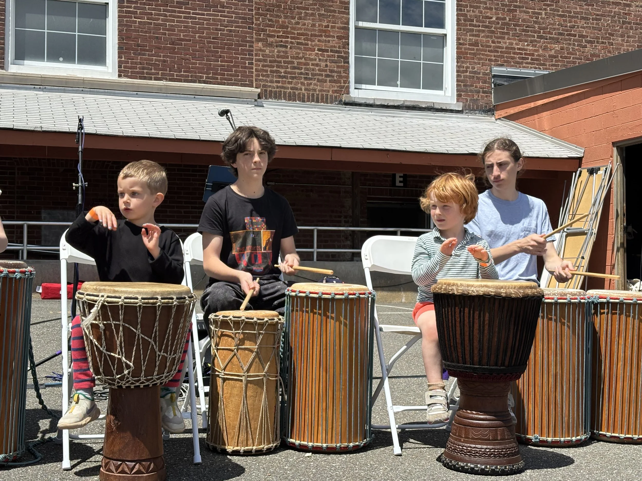 Four children playing African drums outdoors in front of a brick building.