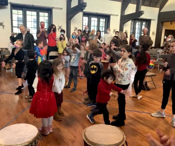 Children and adults gathered in a spacious room with wooden floors, participating in a dance or activity, some wearing masks, with drums at the front.