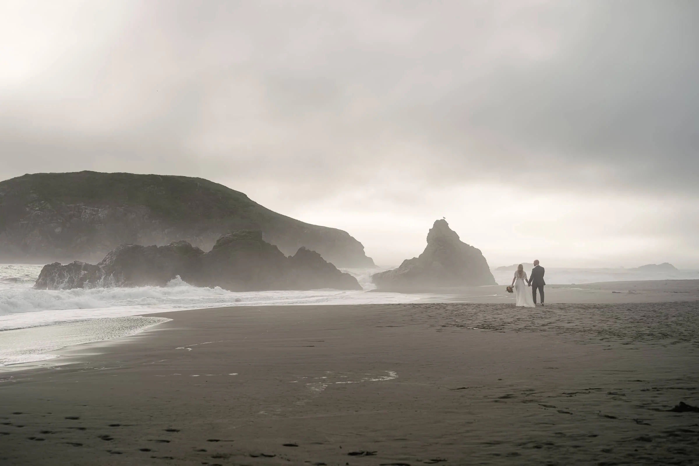 A bride and groom walking hand in hand on a misty Harris beach with large rocks and crashing waves, overcast sky.
