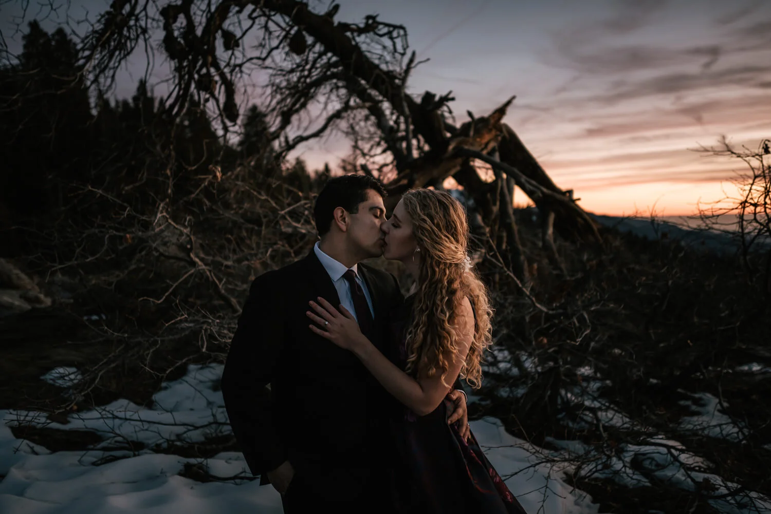 A couple kissing outdoors during sunset in a snowy landscape with fallen tree branches.
