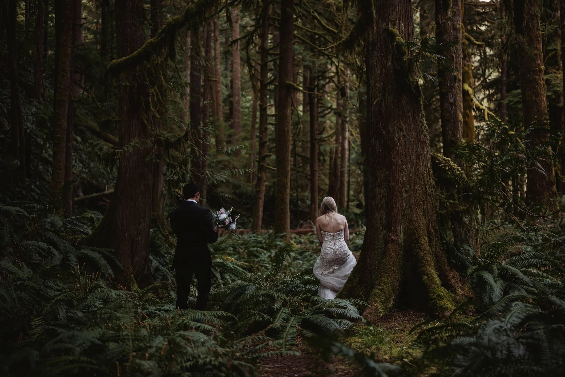 A bride in a white wedding gown and a groom in a black suit are in a dense forest, with tall trees and lush greenery around them.