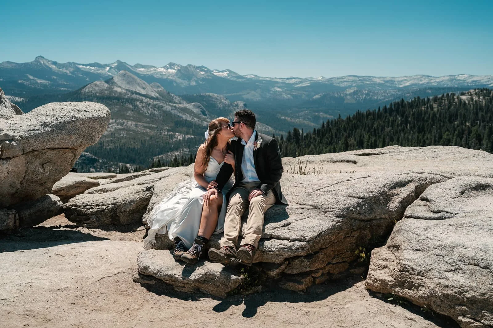 A couple in wedding attire sitting on a large rock, kissing with a mountain range and forest in the background under a clear blue sky.