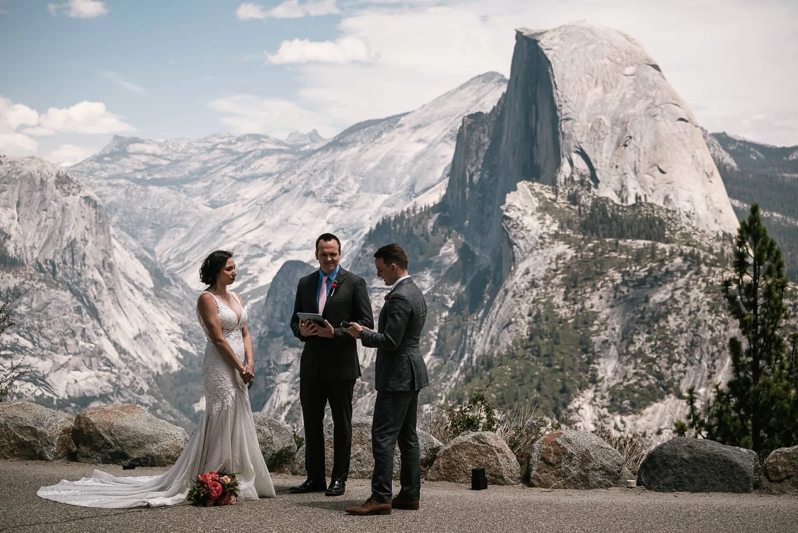 A couple getting married in Yosemite National Park, with mountains and Half Dome in the background, standing with officiant during ceremony.