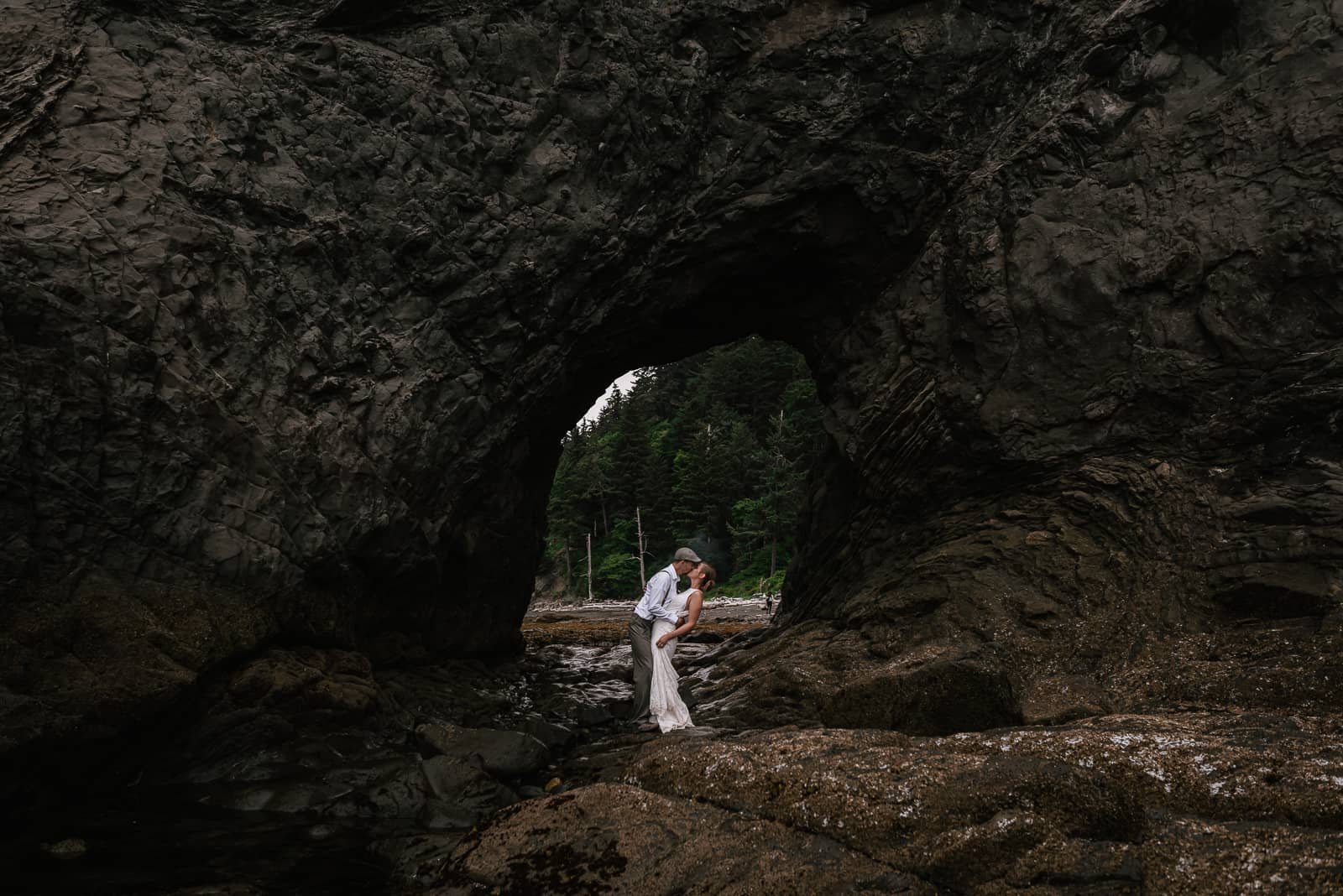 love birds kiss under rock arch at rialto beach during wedding