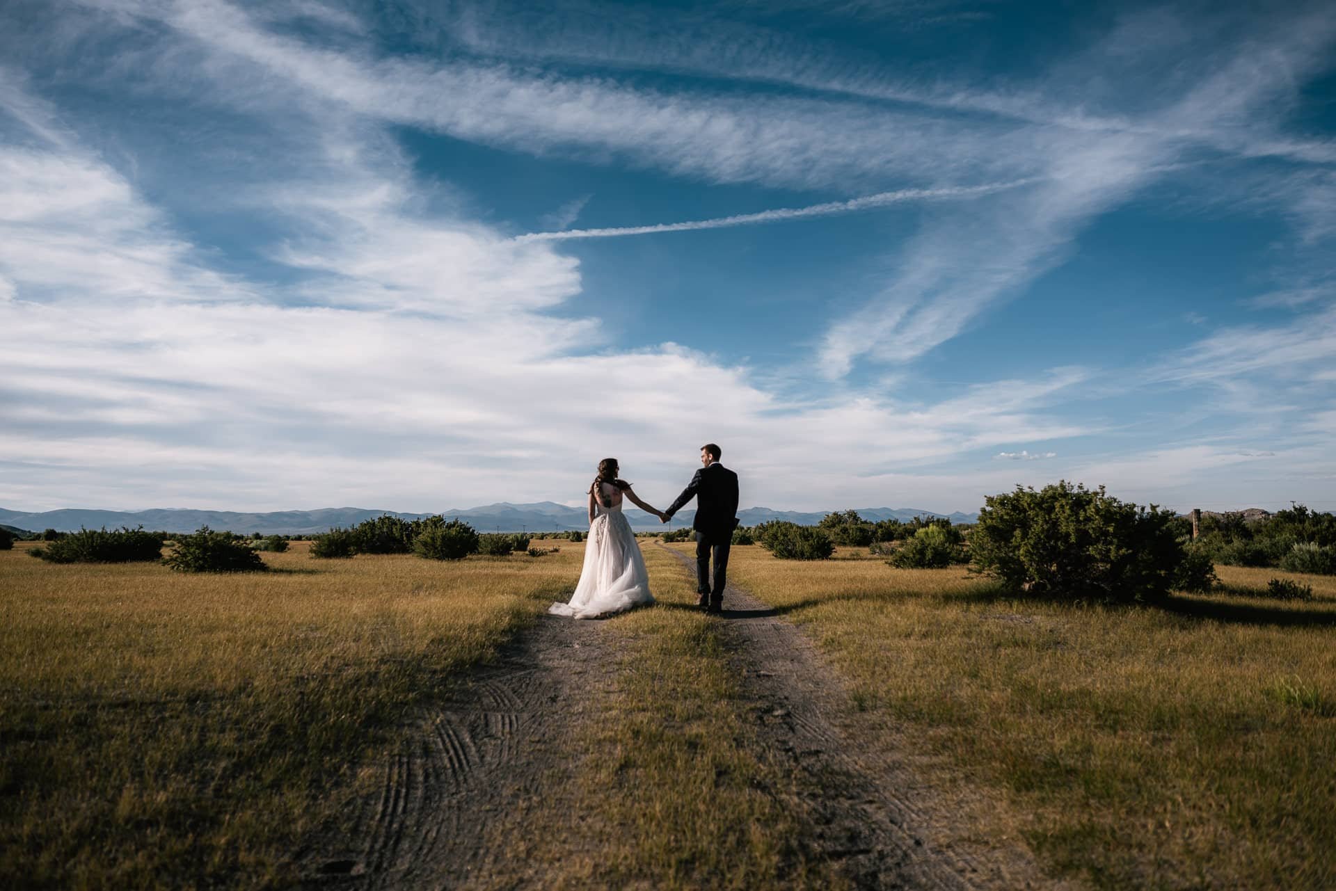 A bride and groom holding hands while walking on a dirt path in an open field with mountains in the distance under a partly cloudy blue sky of the eastern sierra.