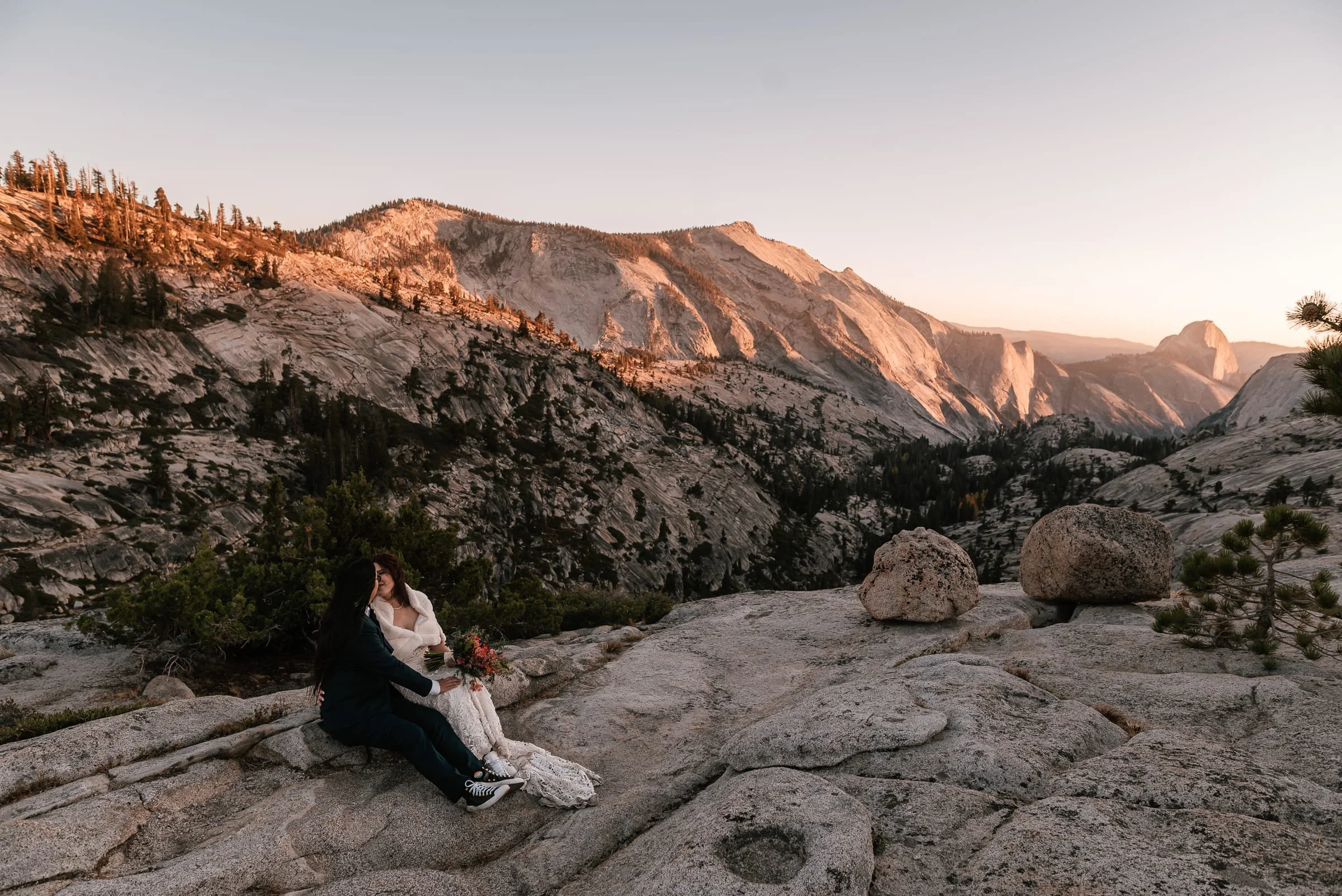 A couple sitting on a rock formation overlooking a mountainous landscape at sunset, with one person holding a bouquet of flowers.
