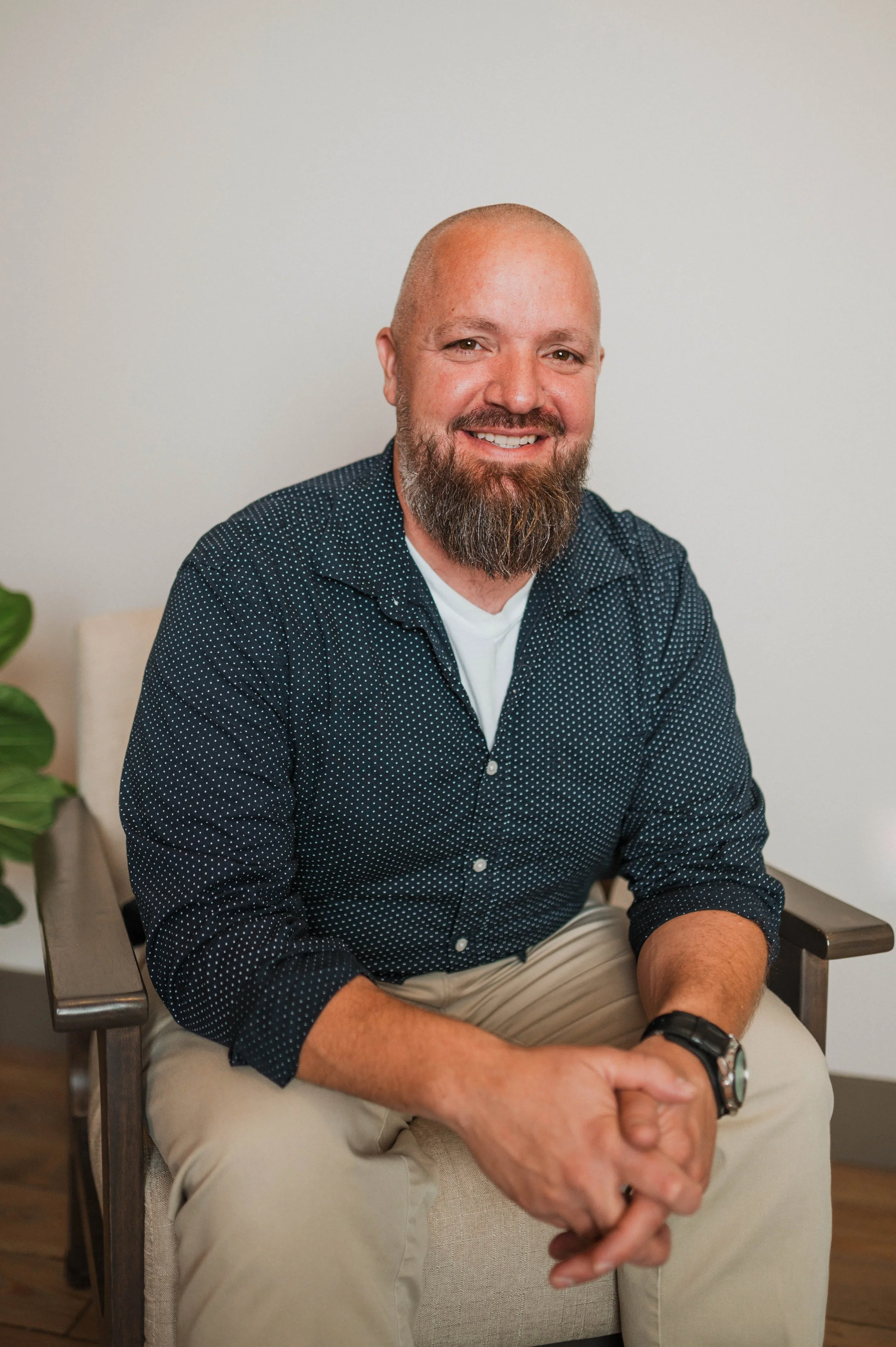 A man with a beard and bald head smiling, sitting in a chair against a plain background, wearing a dark blue polka dot shirt and khaki pants.
