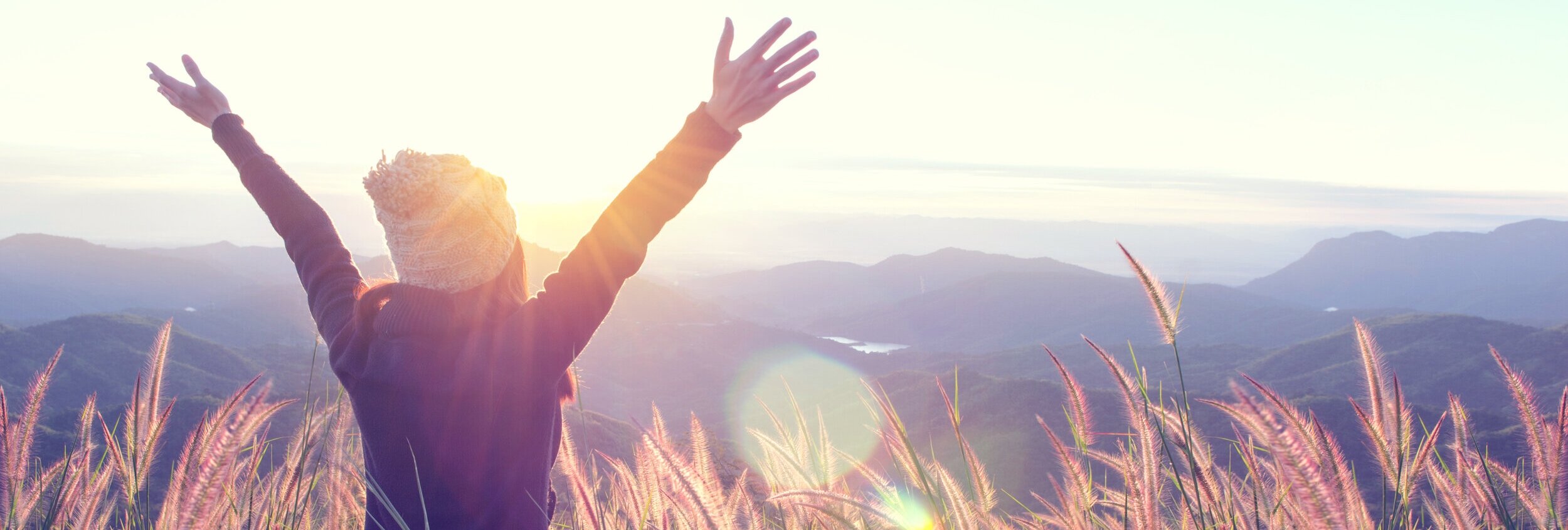 Person standing in a field of tall pink grass with arms raised, overlooking mountains at sunrise or sunset.