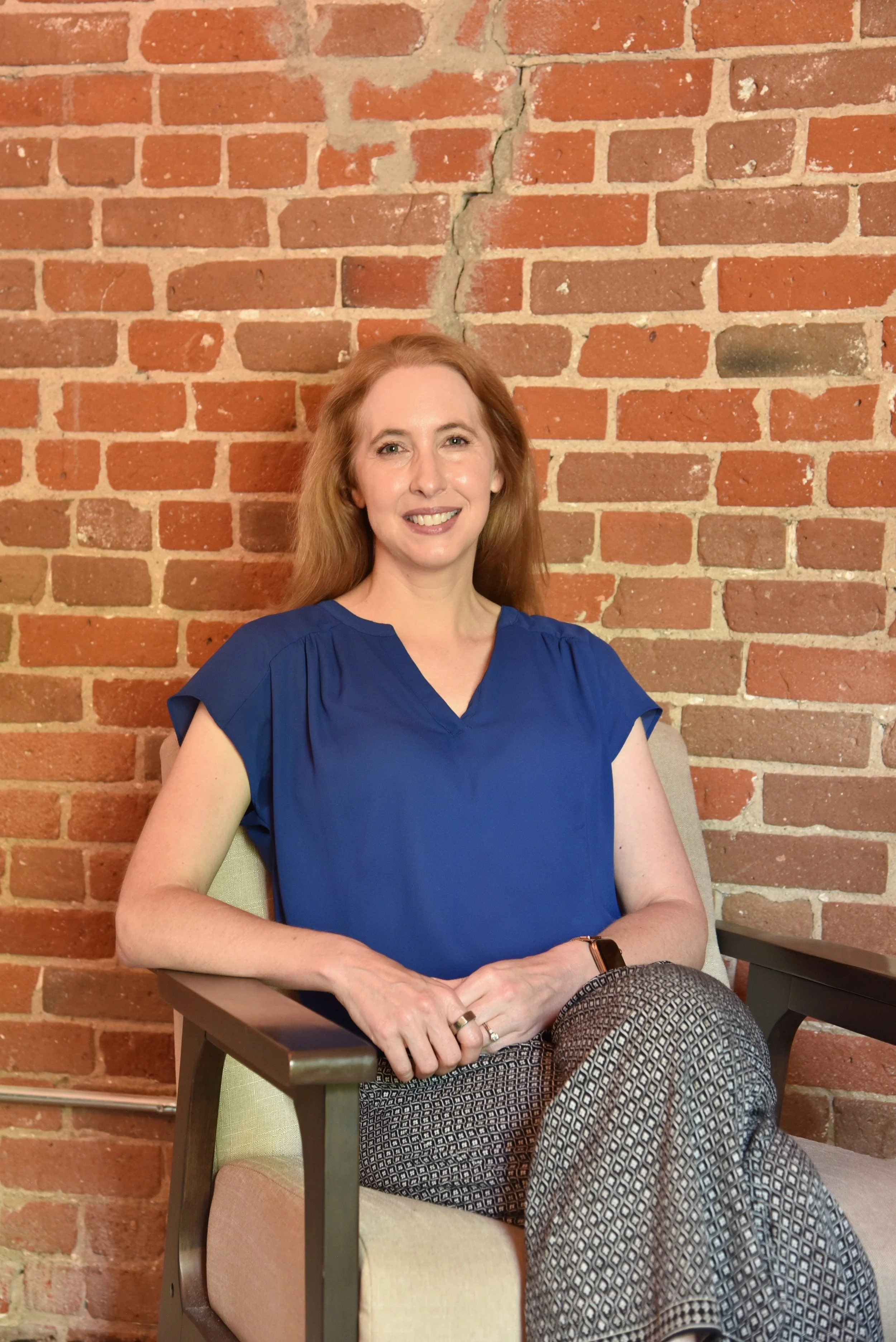 A woman with red hair, smiling, sitting on a beige chair with armrests in front of a brick wall.