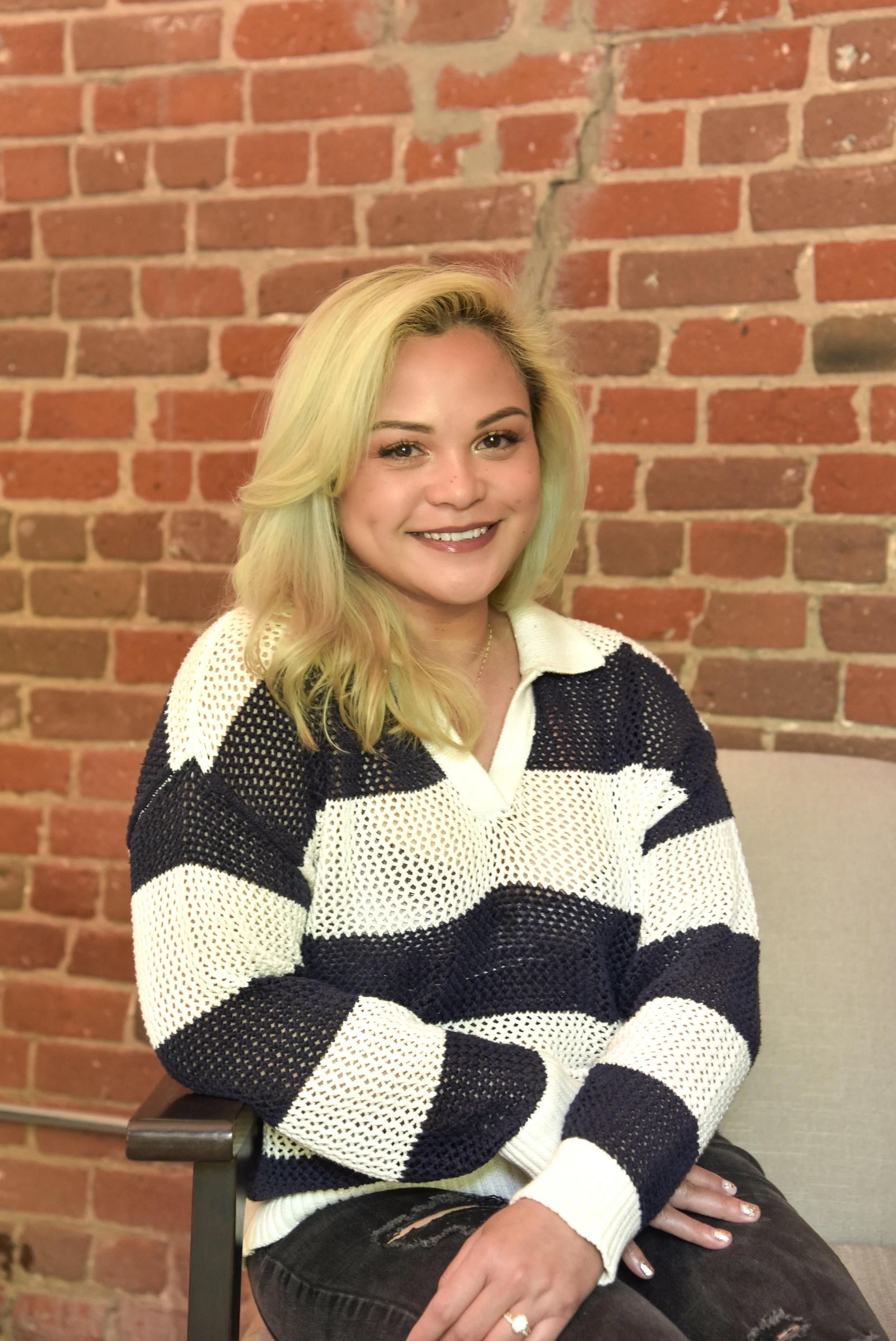 A young woman with blonde hair, wearing a black and white striped sweater, sitting on a beige chair in front of a brick wall, smiling at the camera.