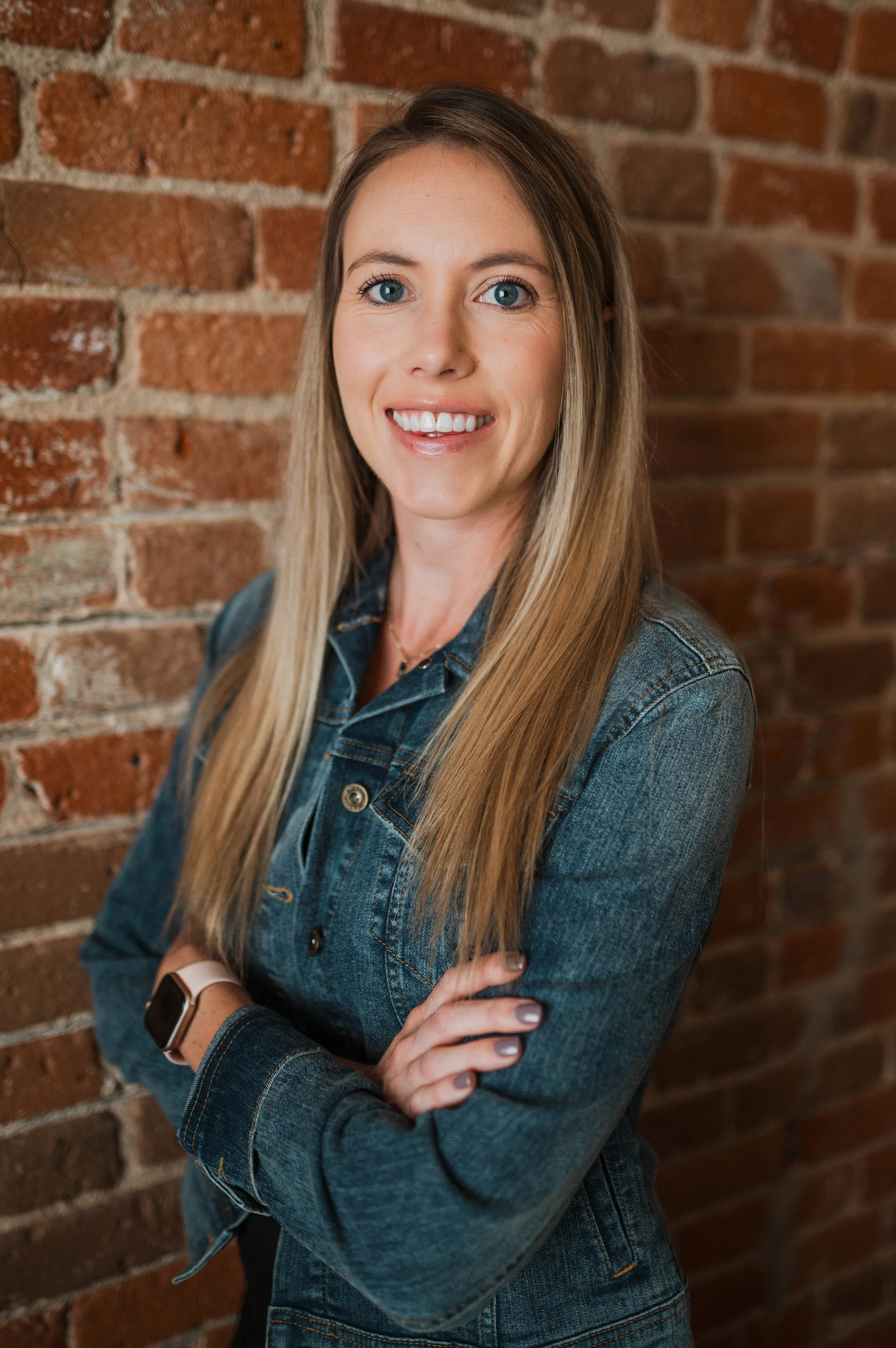 A woman with long blonde hair, blue eyes, and a smile, standing with arms crossed in front of a brick wall, wearing a denim jacket and a smartwatch.