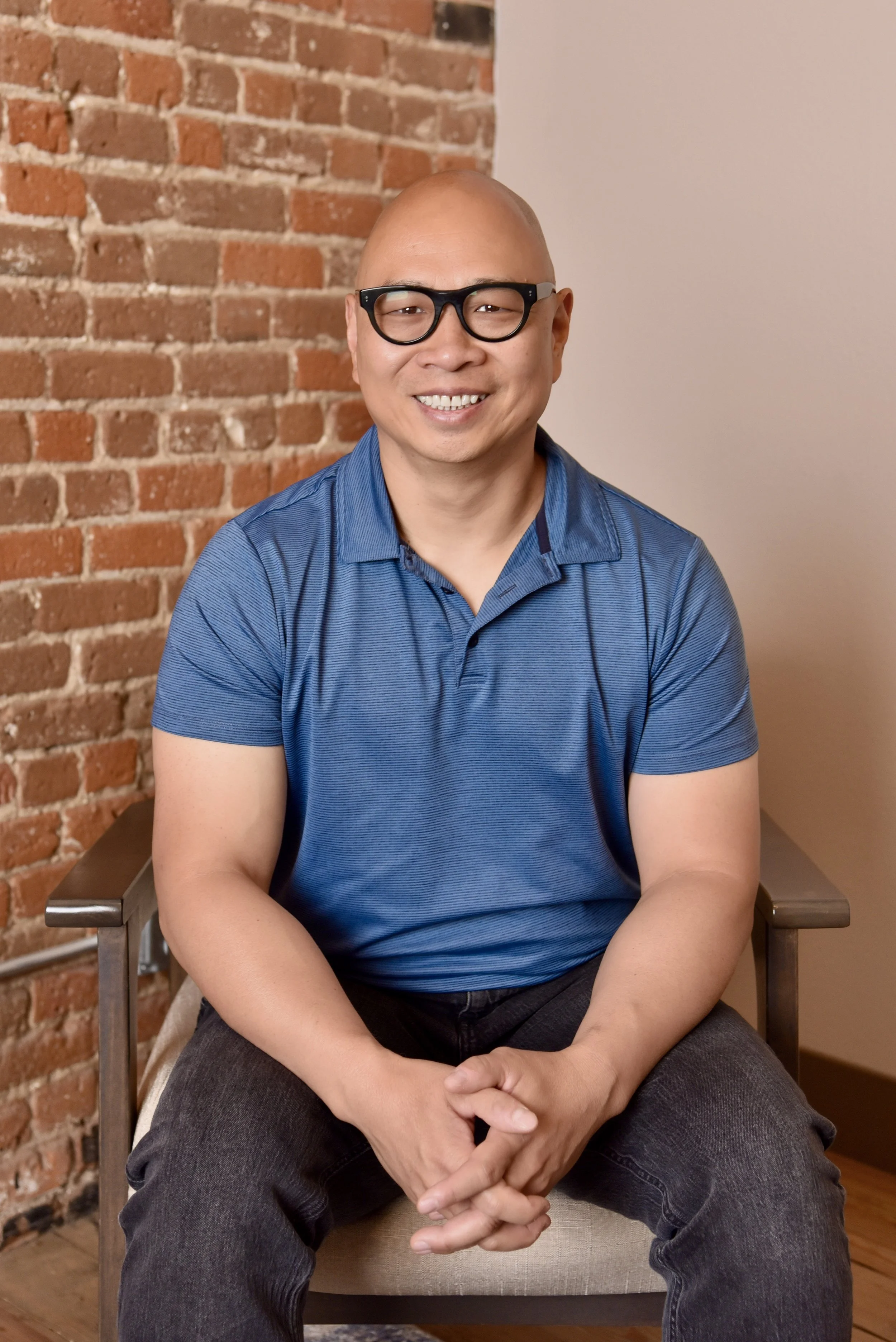 A smiling bald man with glasses wearing a blue polo shirt and black jeans, sitting on a beige chair with a brick wall and a white wall in the background.