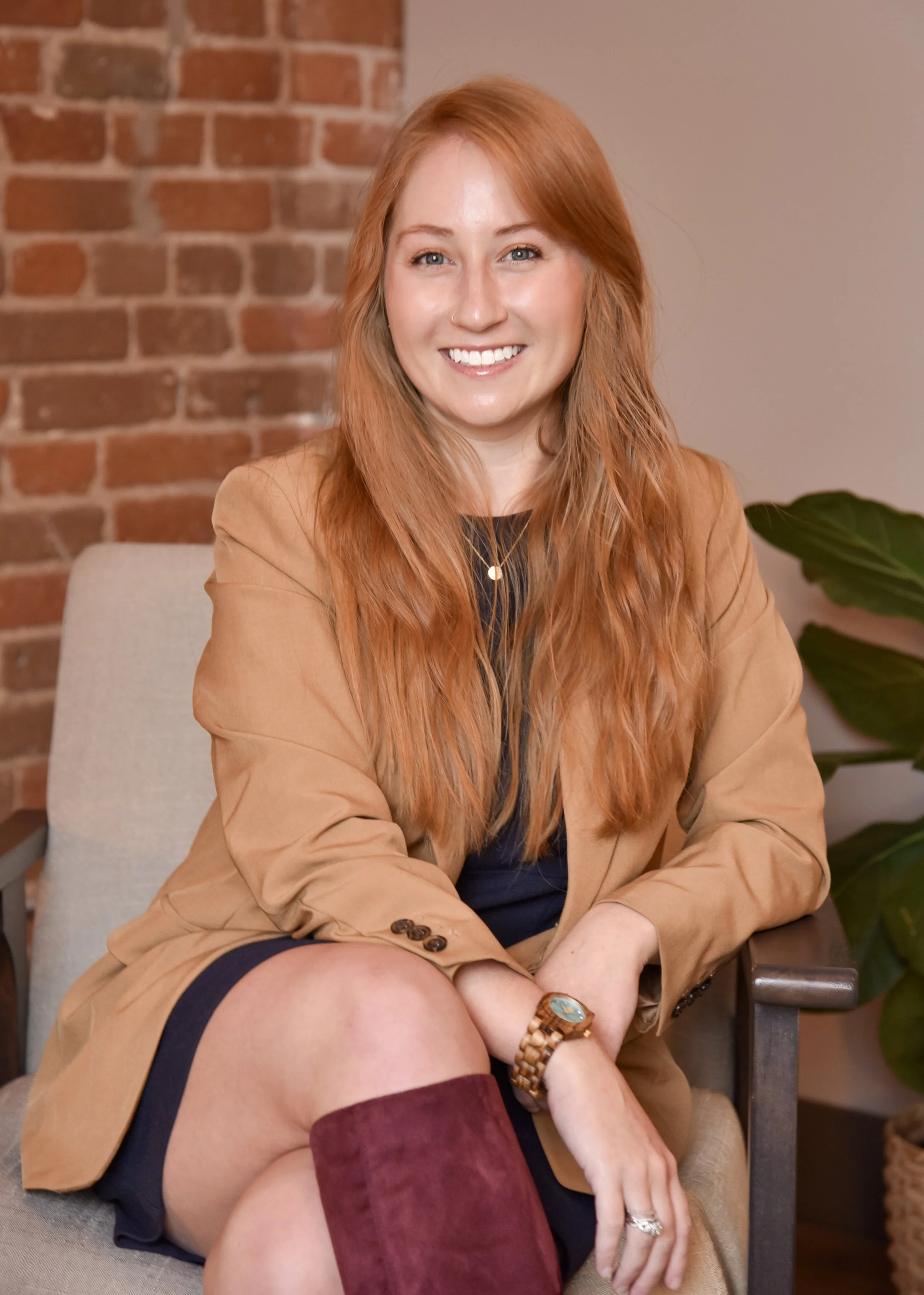 A woman with long red hair sitting on a beige chair in front of a brick wall, smiling at the camera.