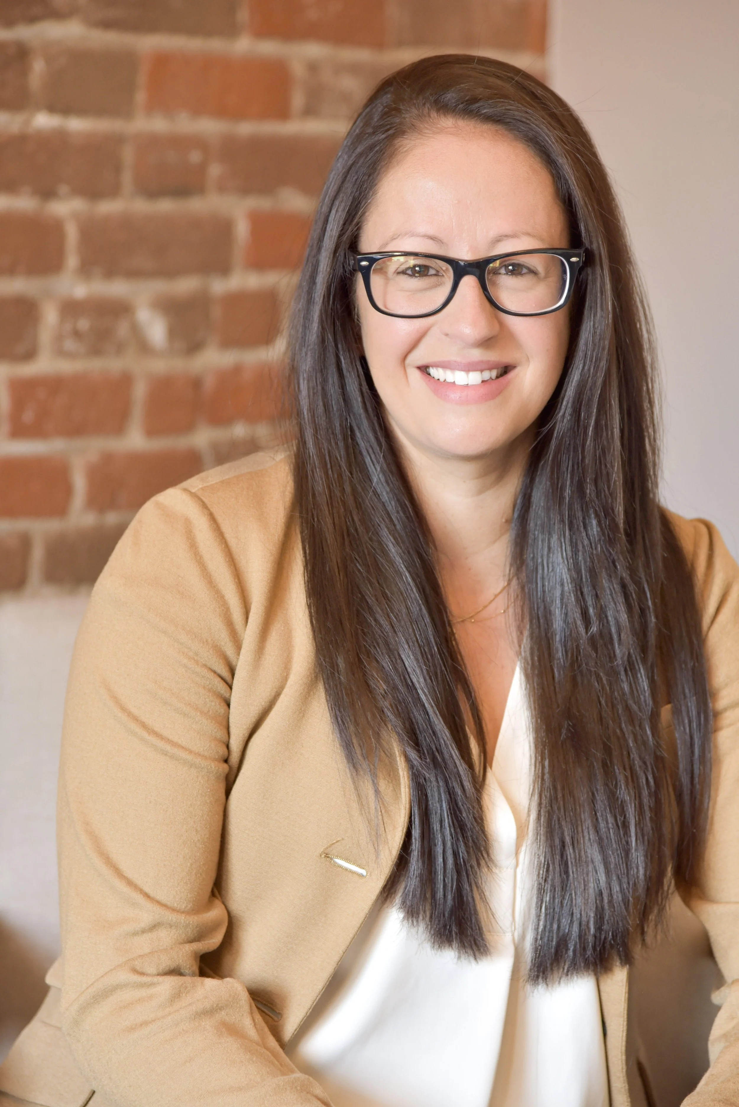 A woman with long dark hair, glasses, and a beige blazer smiling at the camera, sitting against a brick wall.