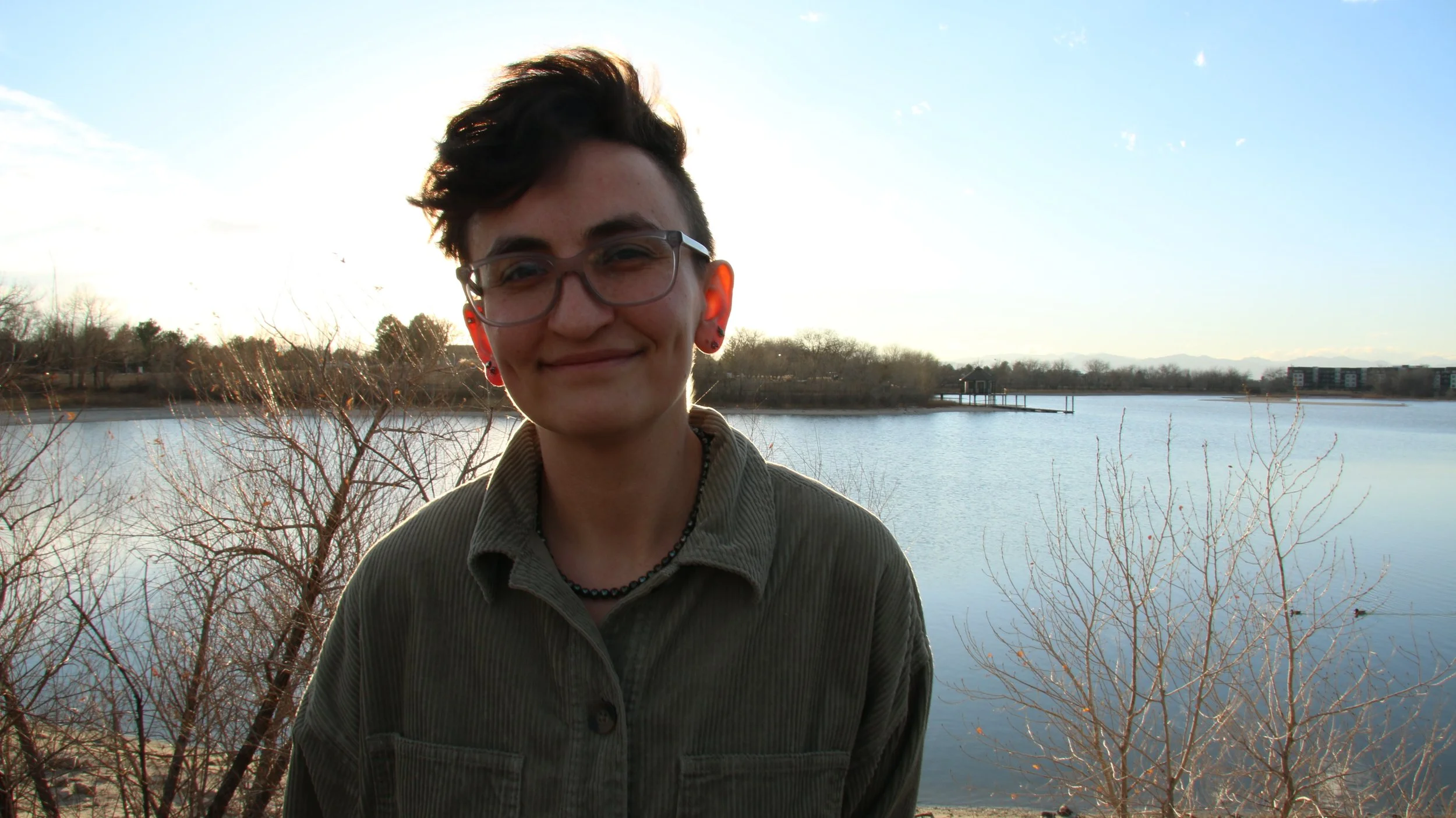 Young person with short dark hair, glasses, and earrings smiling outdoors near a lake with leafless bushes and trees, under a partly cloudy sky.