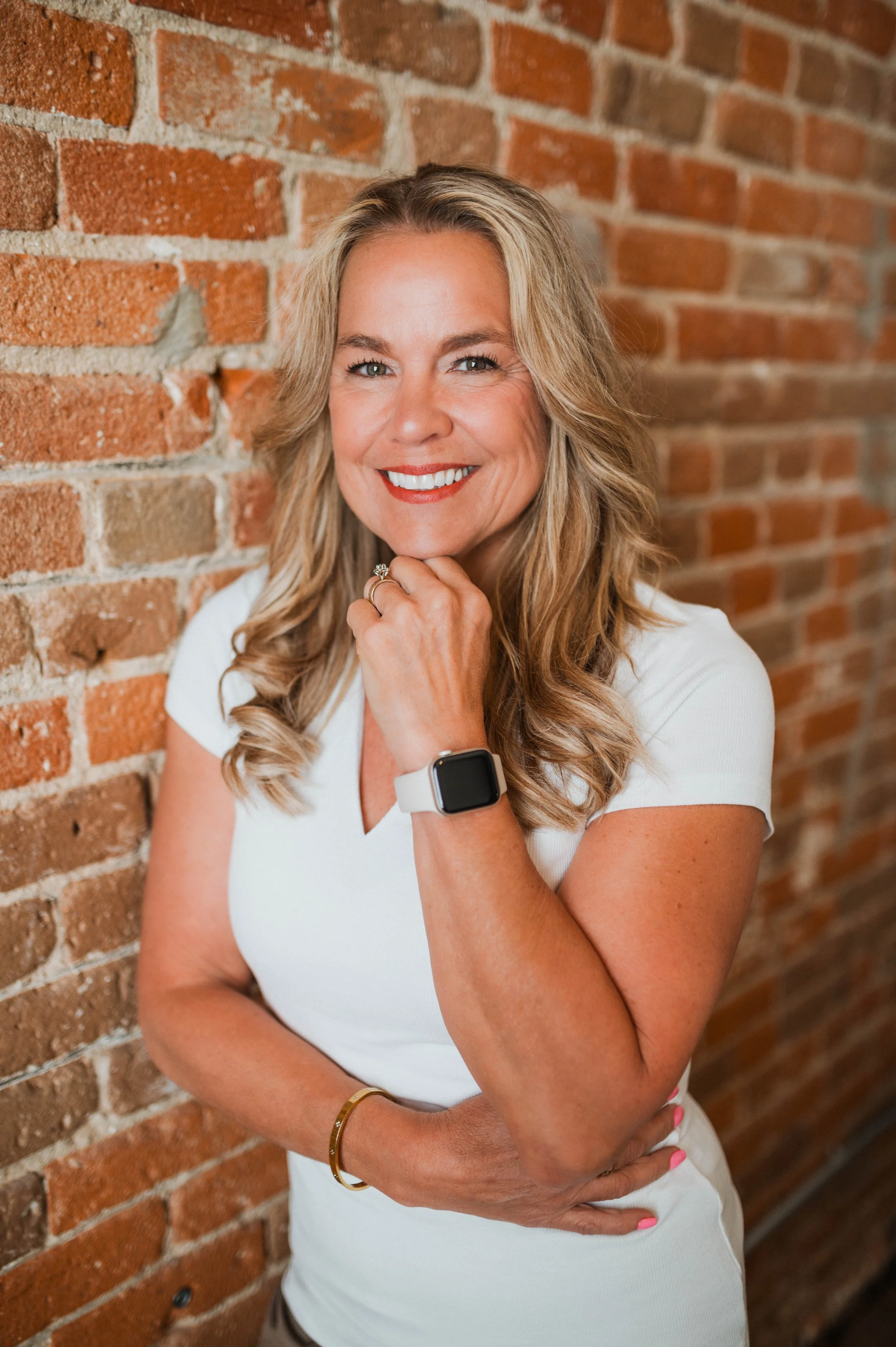 A woman with long blonde hair and a white shirt, smiling and resting her chin on her hand, standing against a brick wall.