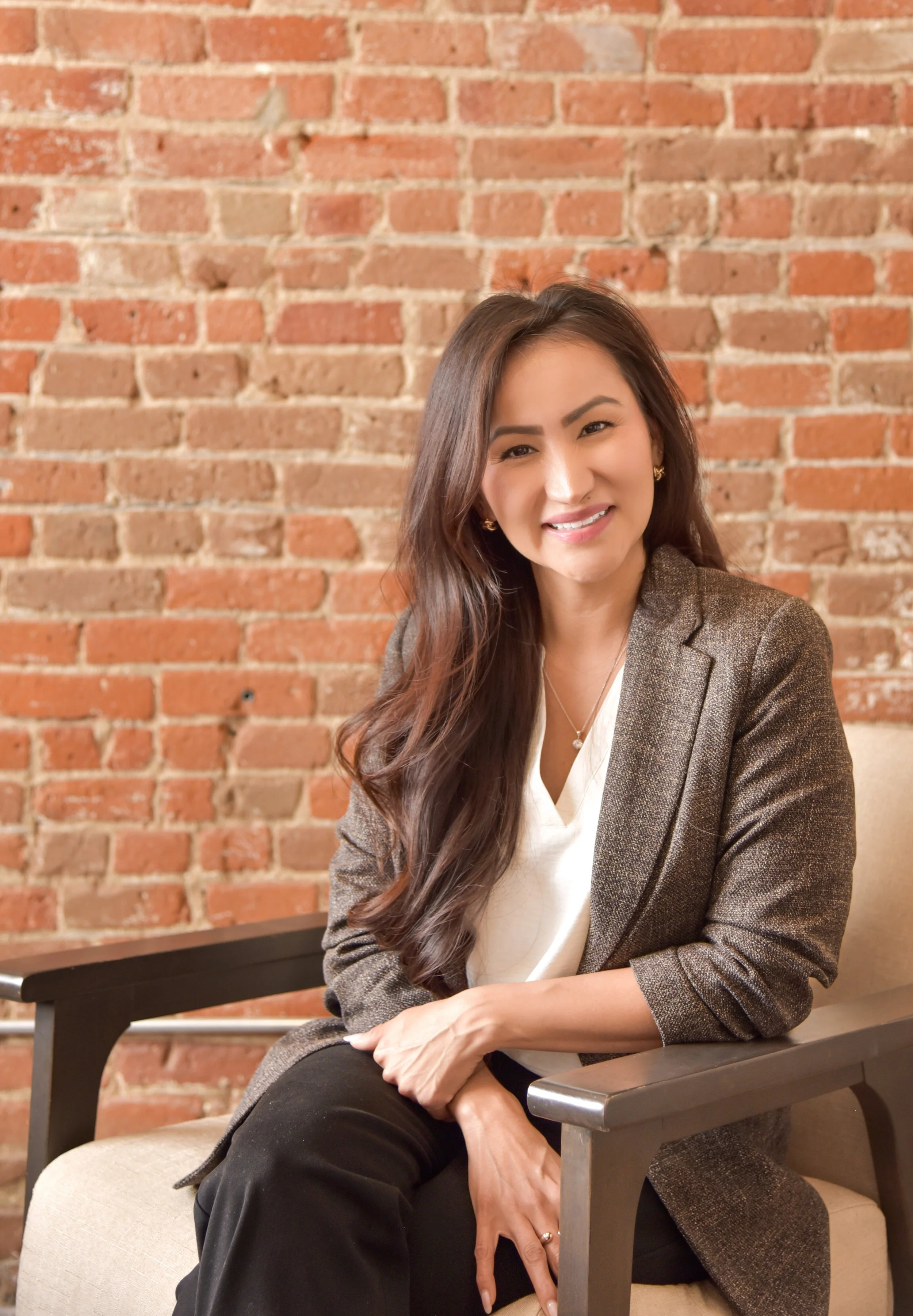 A woman with long, wavy brown hair, smiling and wearing a brown blazer, white blouse, and black pants, sitting on a beige chair with wooden armrests in front of a brick wall.