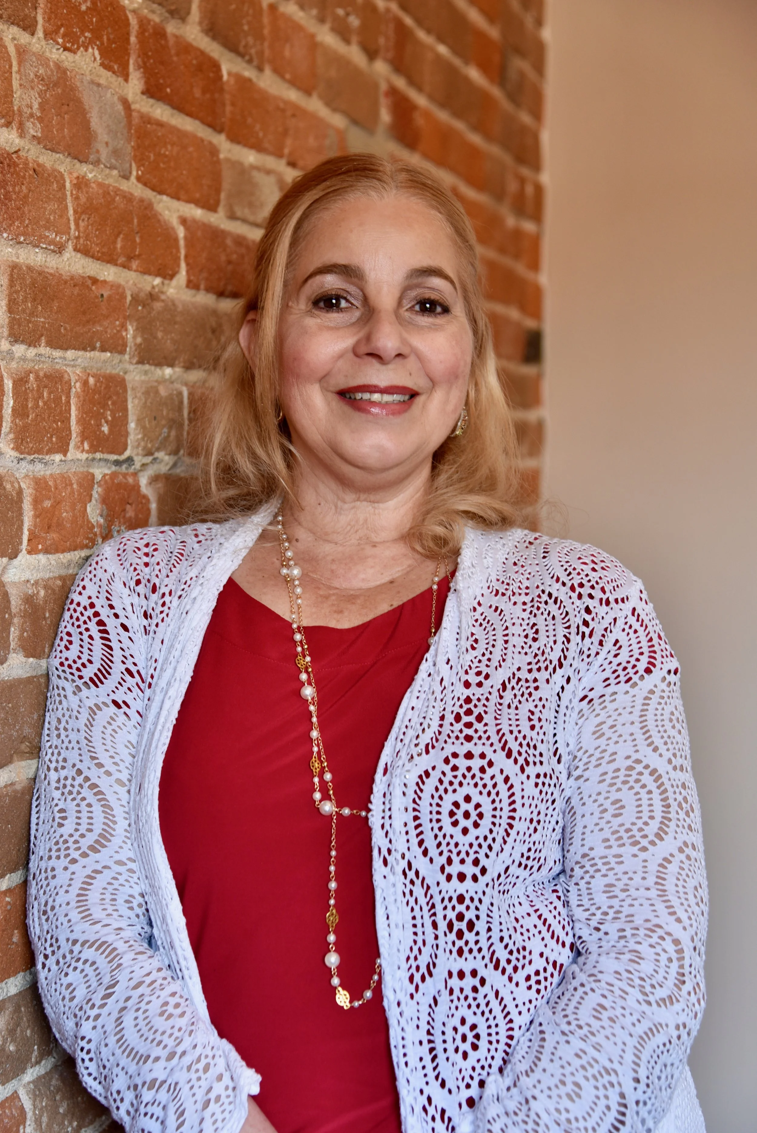 A smiling middle-aged woman with blonde hair, wearing a white lace cardigan over a red top, gold jewelry, standing against an exposed brick wall.