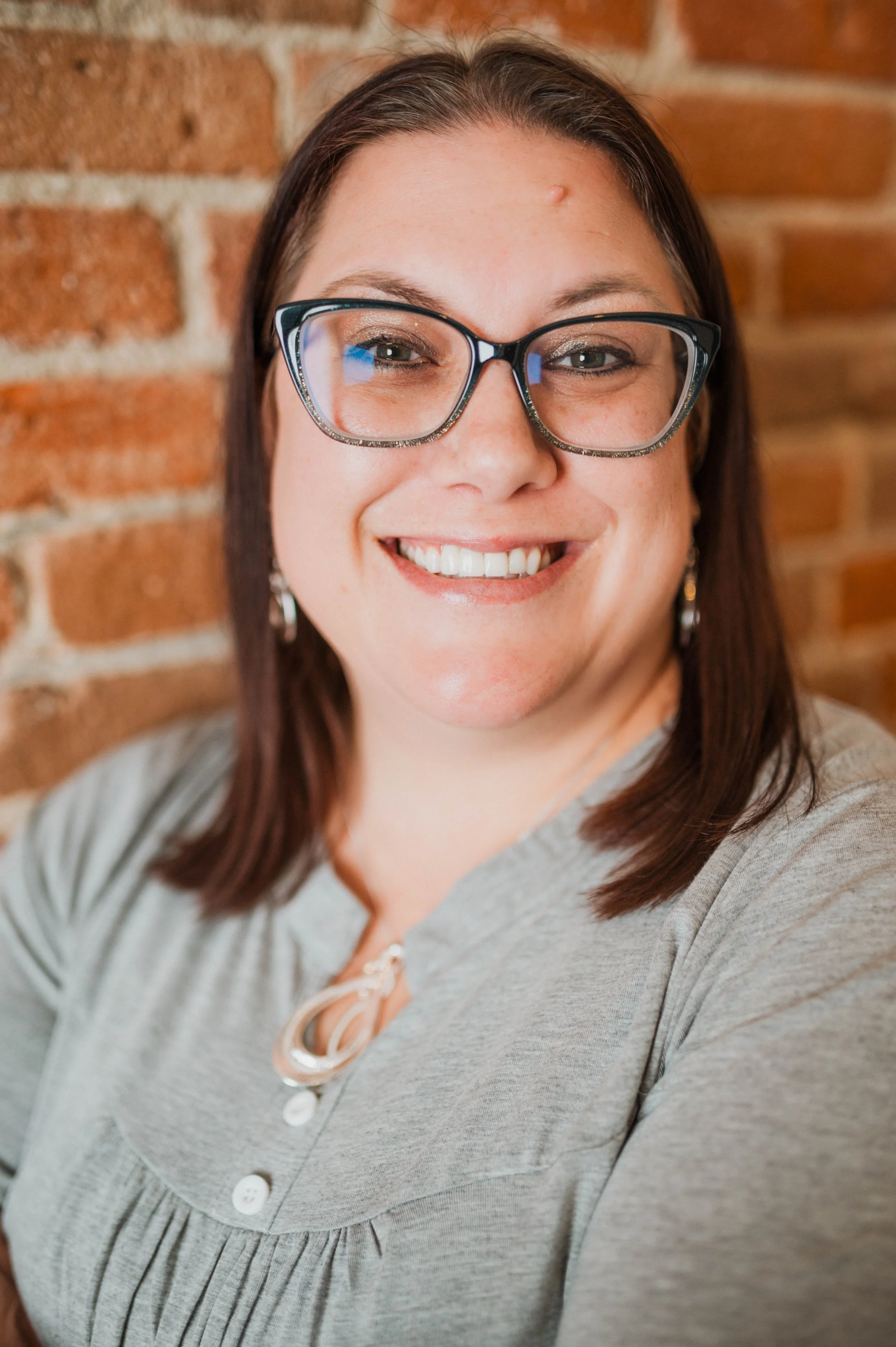 A smiling woman with glasses and earrings, wearing a gray top, standing in front of a brick wall.