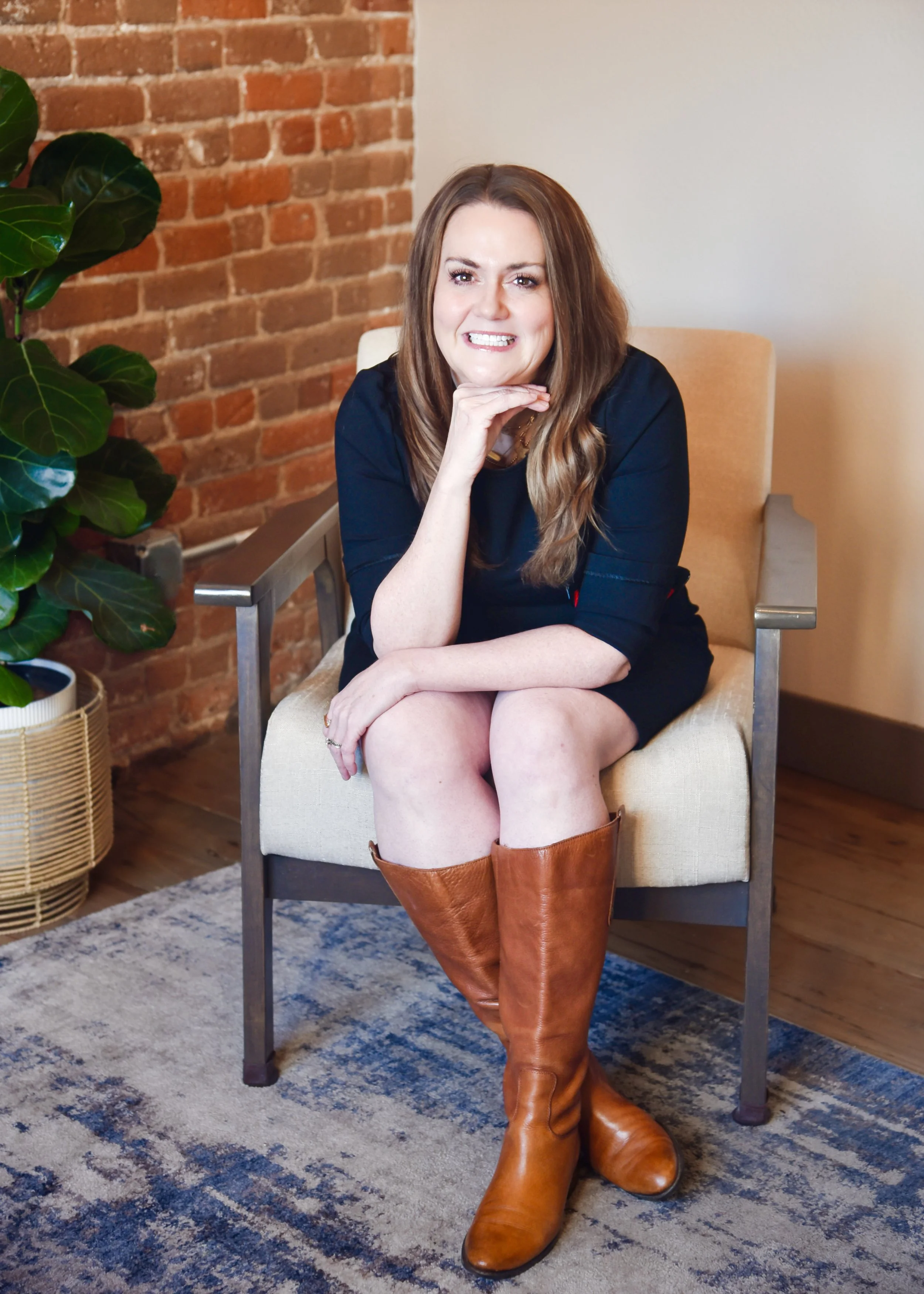 A woman with long brown hair, wearing a black dress and brown knee-high boots, is sitting on a beige armchair with her legs crossed. She is smiling, resting her chin on her right hand, in a room with a brick wall, a houseplant, and a rug.