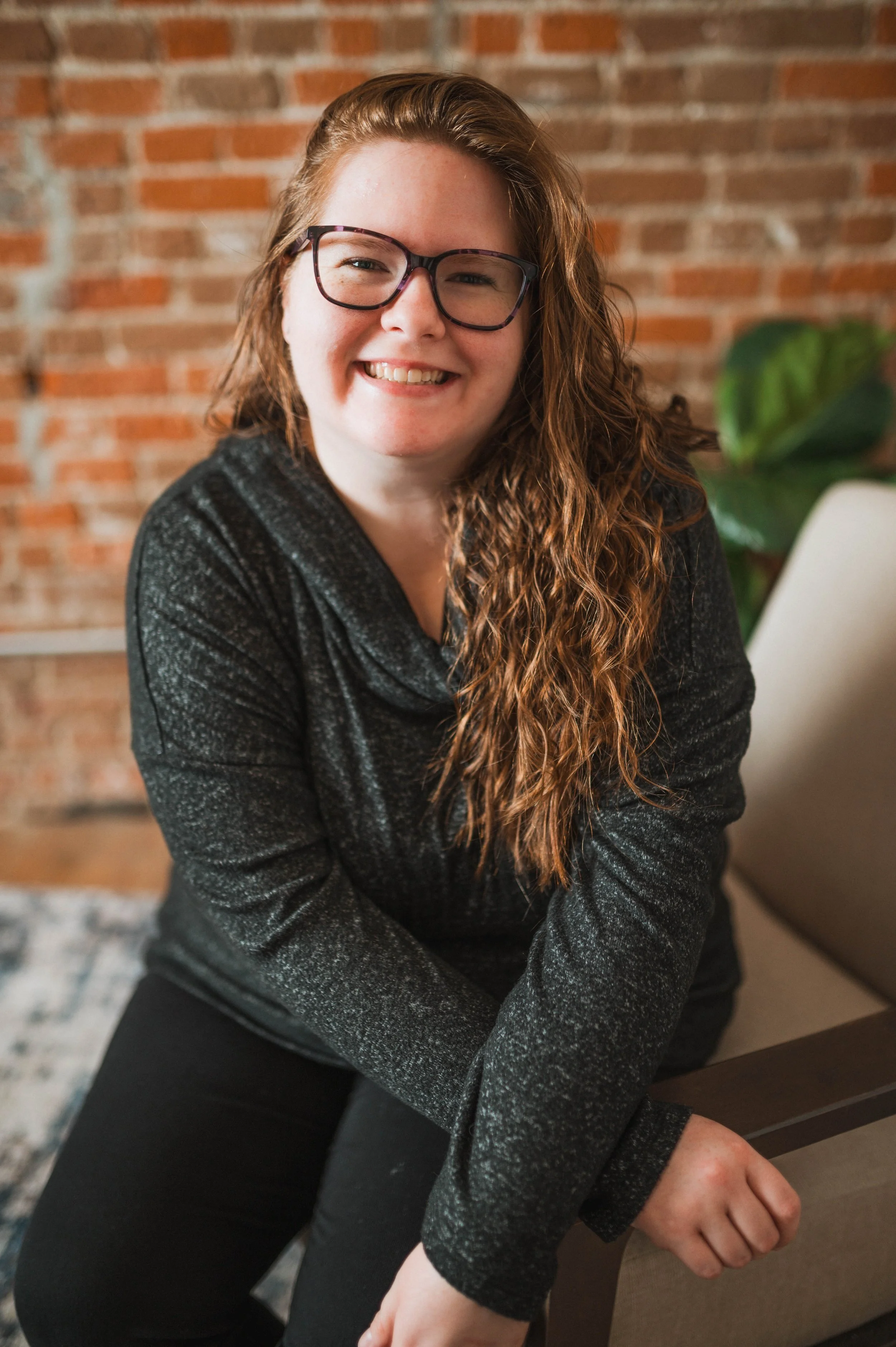 A young woman with curly red hair, glasses, and a black long-sleeve top sitting on a chair, smiling at the camera in a room with a brick wall and some green plants.