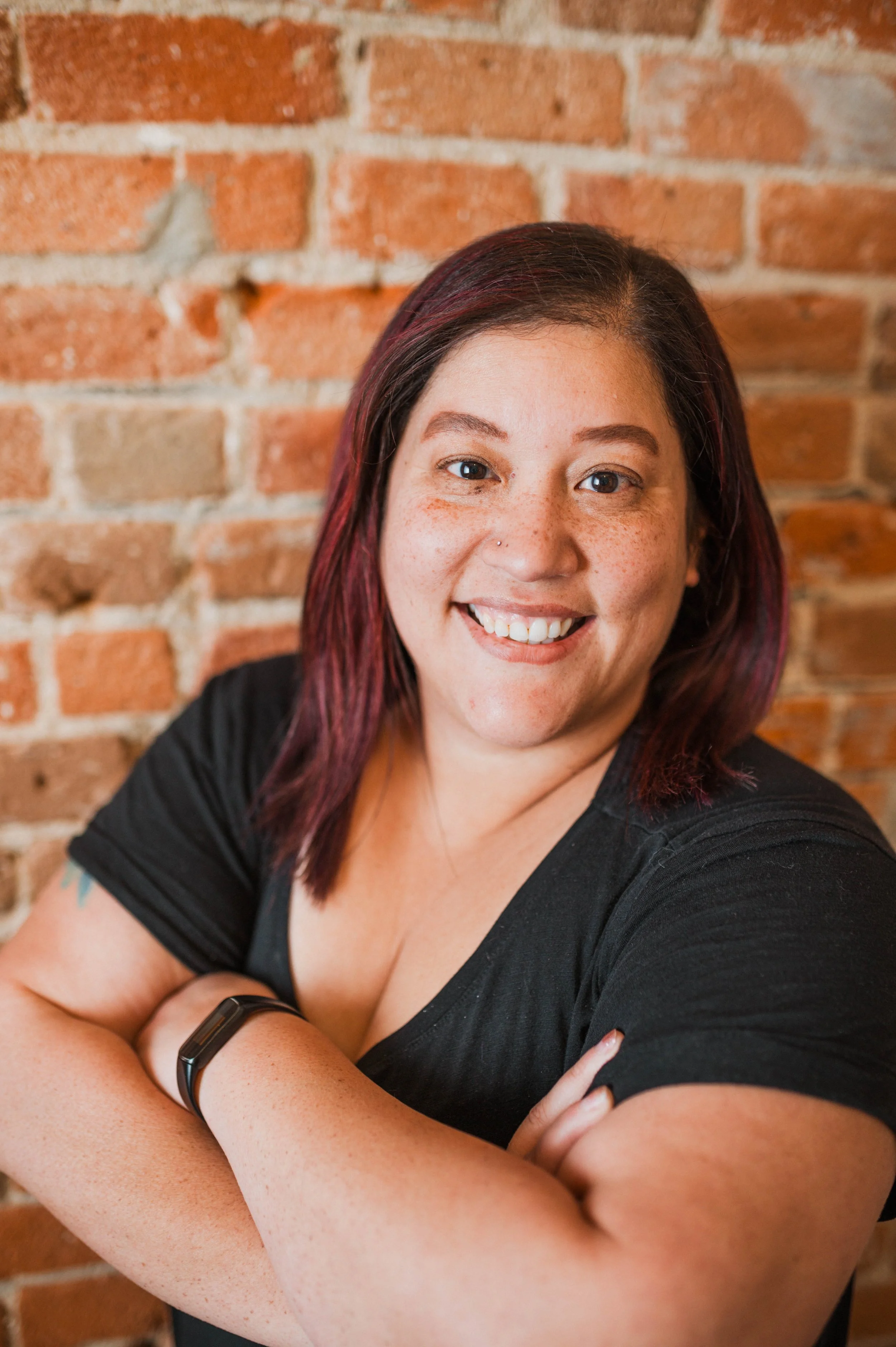 A woman with shoulder-length purple hair, smiling confidently with your arms crossed, against a brick wall background.