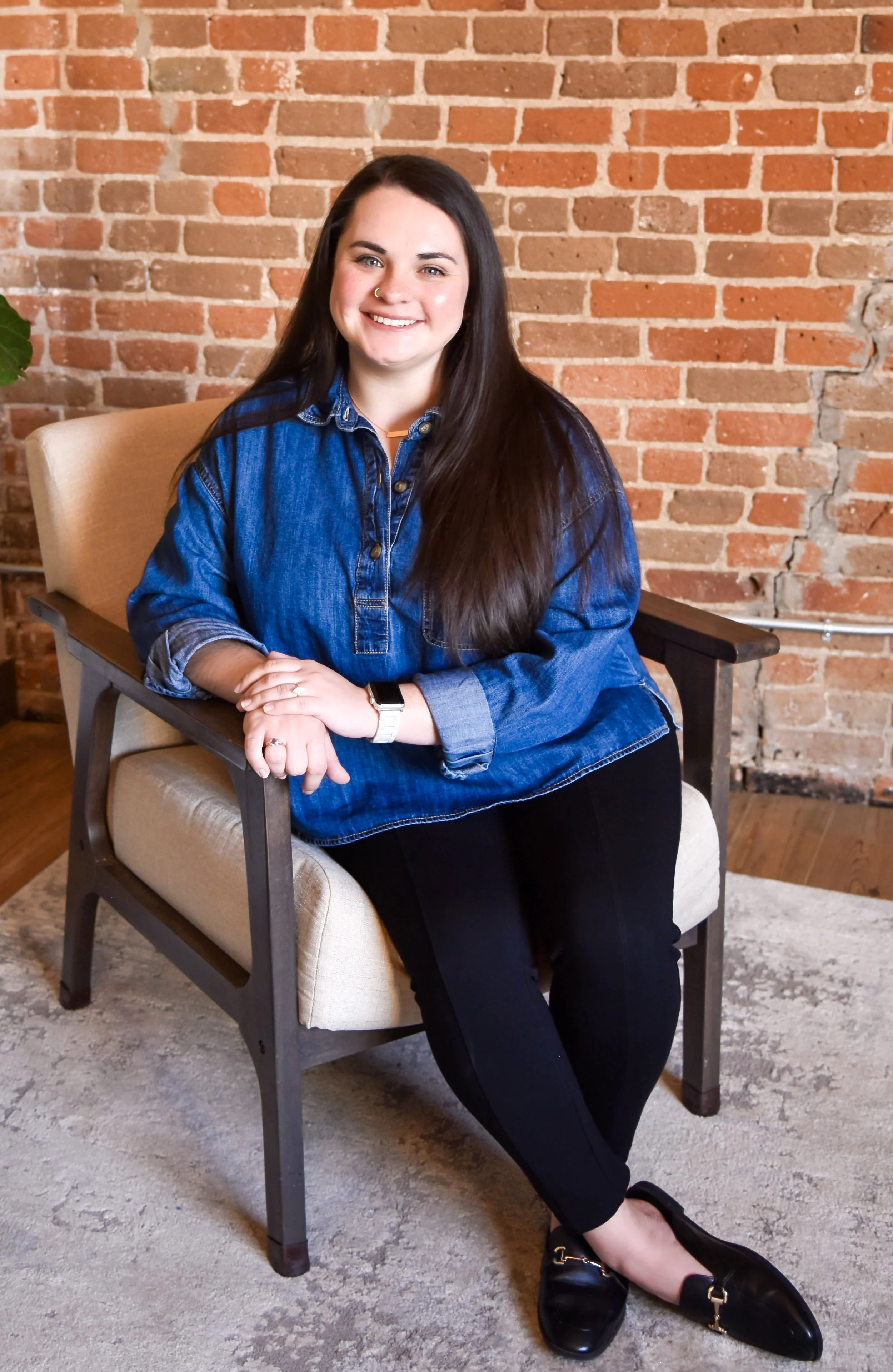A young woman sitting on a beige armchair with a brick wall background, smiling, wearing a denim jacket and black pants, with a large green plant to the left.