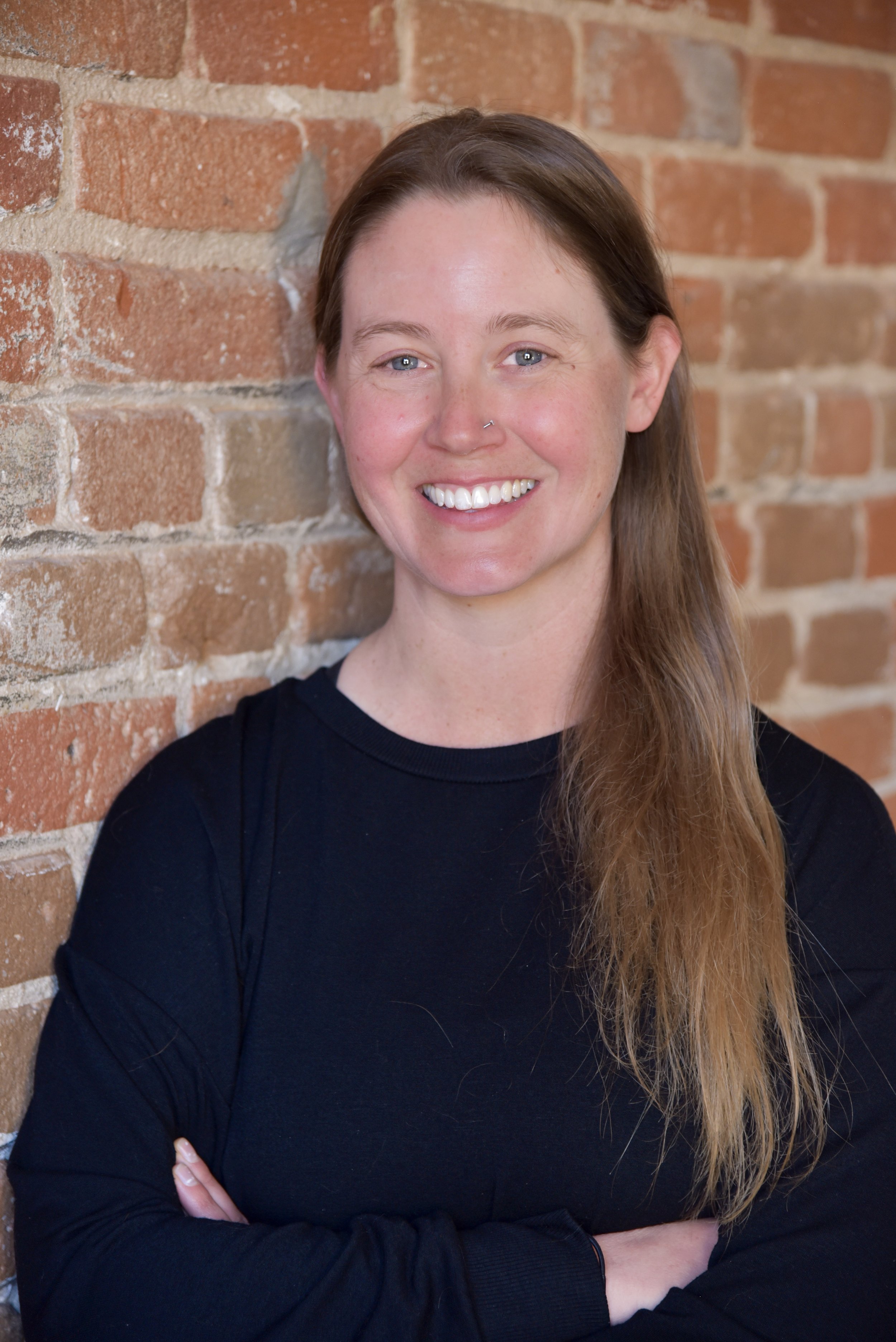 A woman with long brown hair and a nose piercing, smiling and wearing a black long-sleeve top, standing against a brick wall.