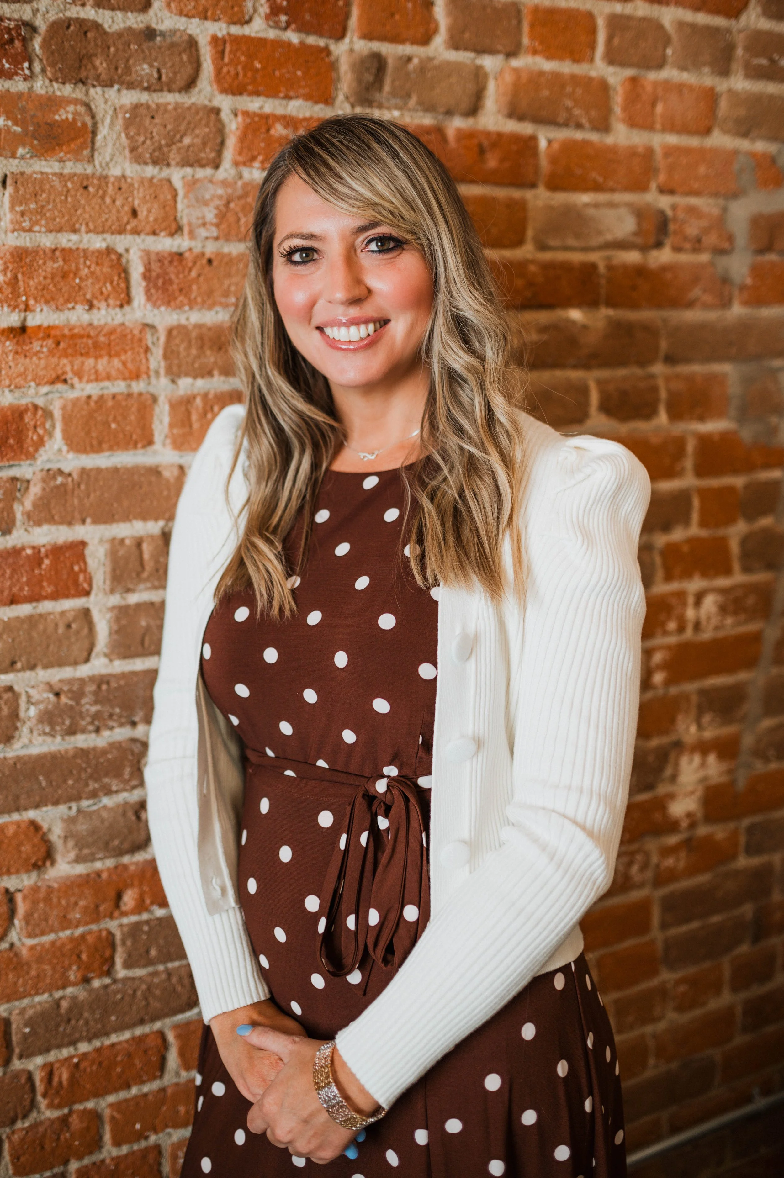 A woman with long blonde wavy hair and light skin standing in front of a brick wall, smiling at the camera, wearing a brown dress with white polka dots, a cream-colored cardigan, a bracelet, and a necklace.