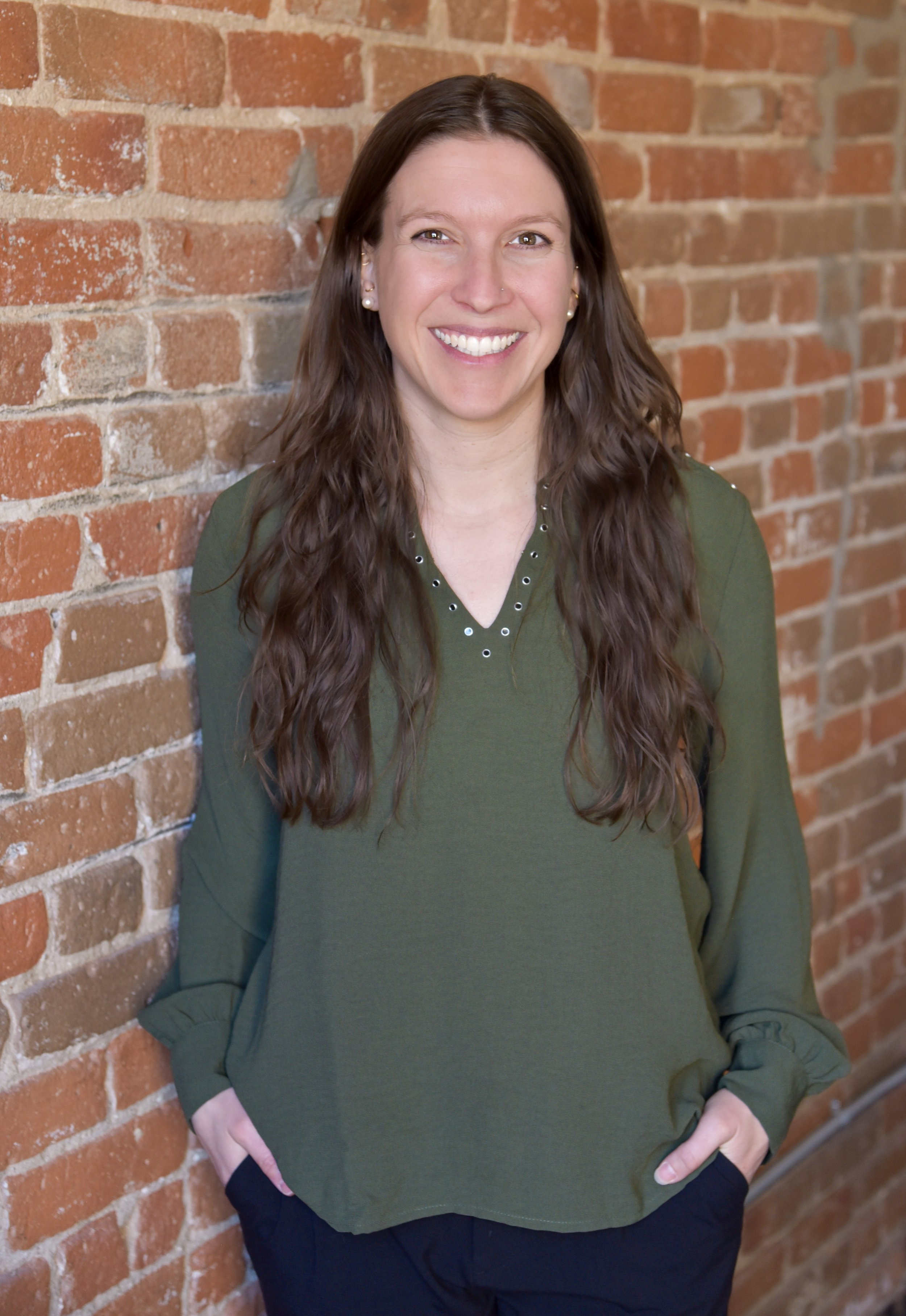 Woman with long brown hair, wearing a green shirt, standing in front of a brick wall with her hands in her pockets. 