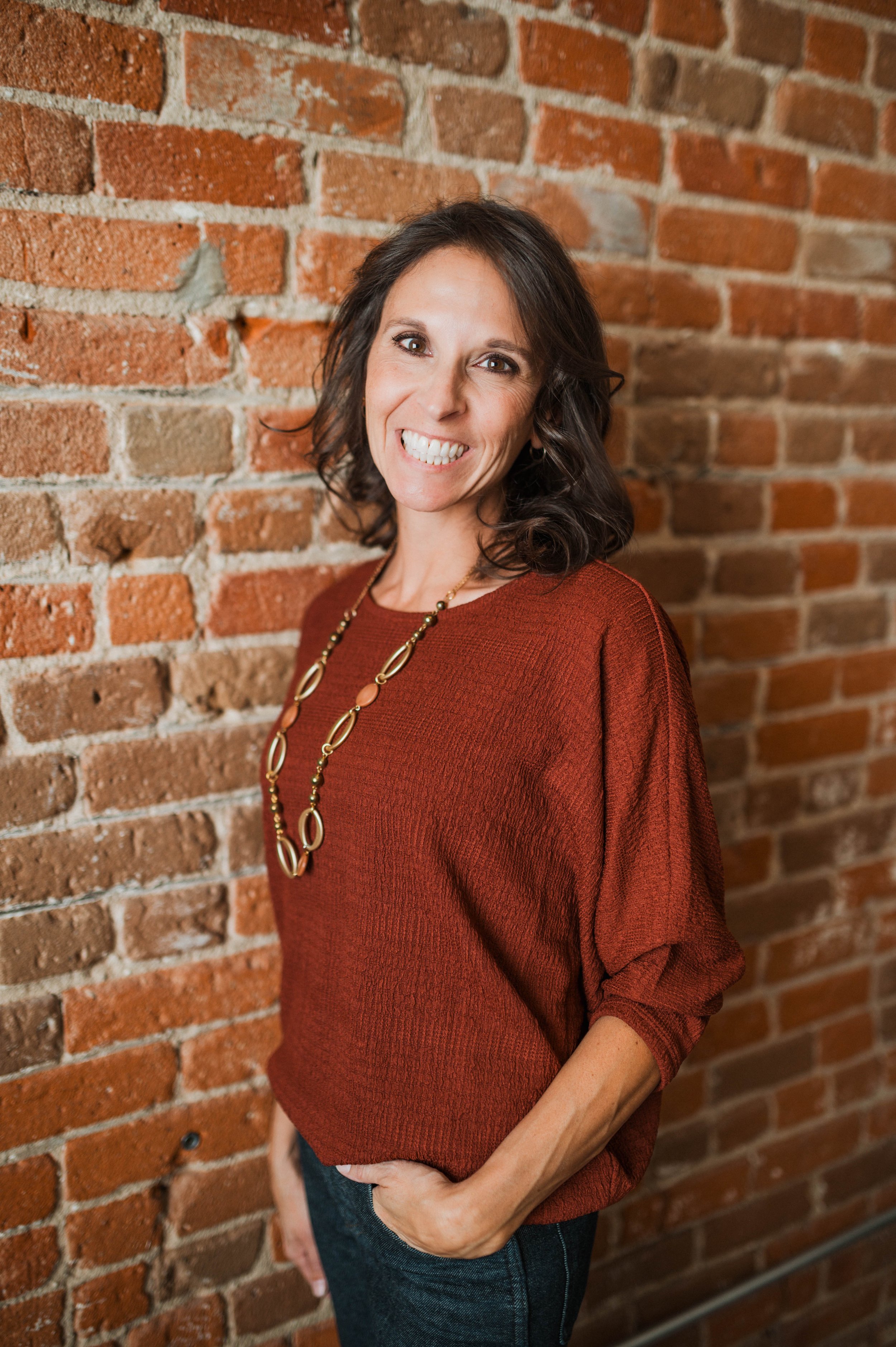 A woman with shoulder-length dark hair, smiling, wearing a rust-colored sweater and a gold necklace, standing against a brick wall.