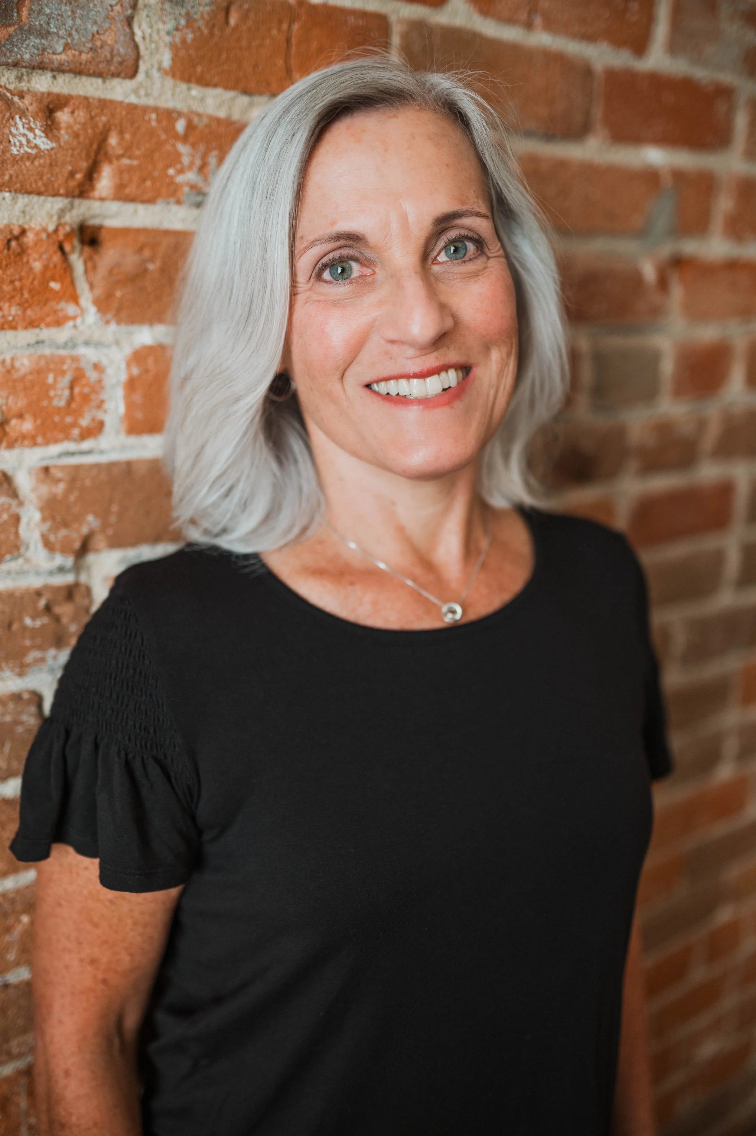 A middle-aged woman with shoulder-length gray hair, blue eyes, and freckles, smiling while leaning against a red brick wall, wearing a black short-sleeve top and a silver necklace.