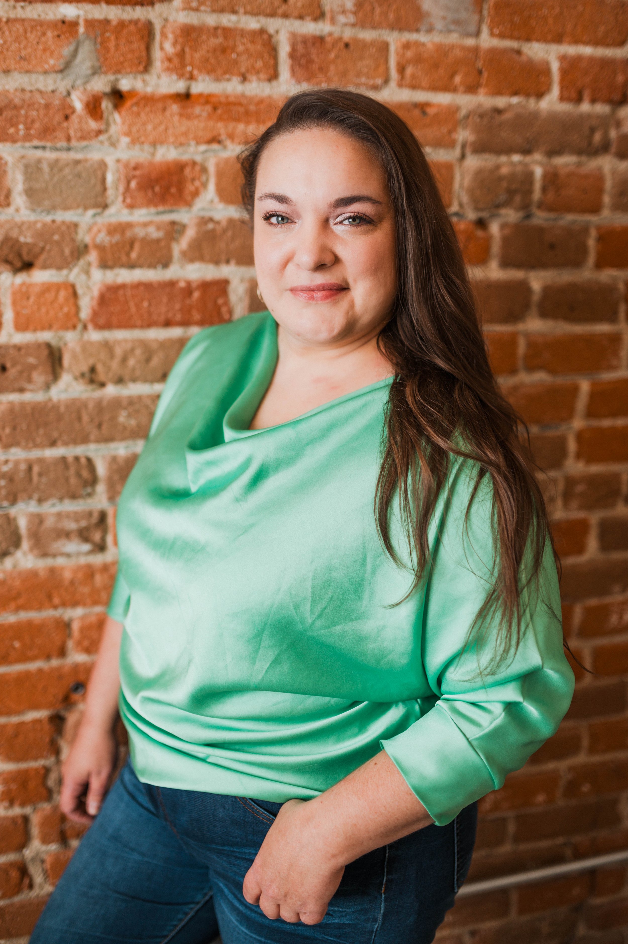 Woman with long brown hair wearing a green satin blouse and blue jeans standing in front of an exposed brick wall.