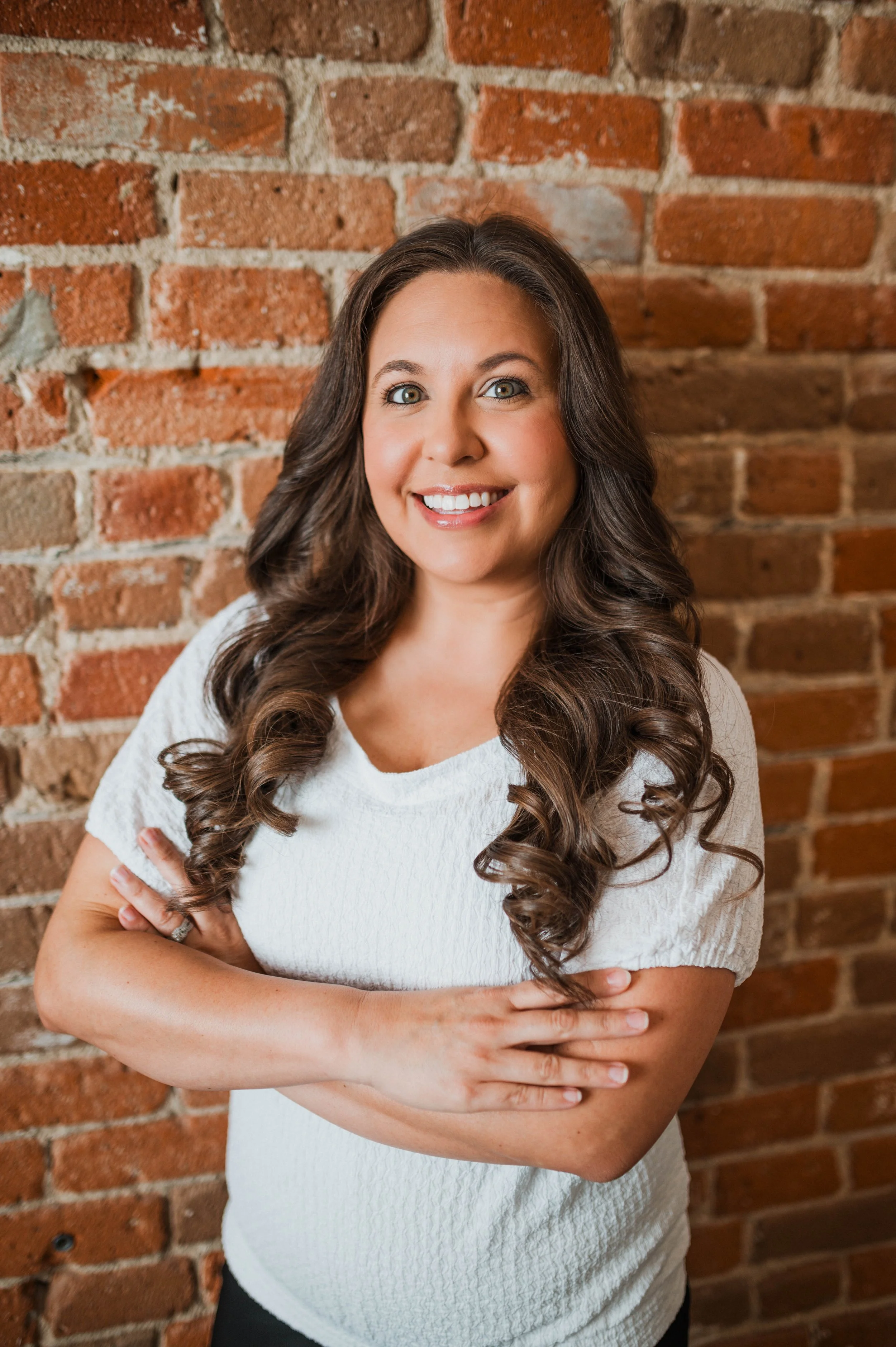 A woman with long, curly brown hair standing in front of a brick wall, smiling and crossing her arms.
