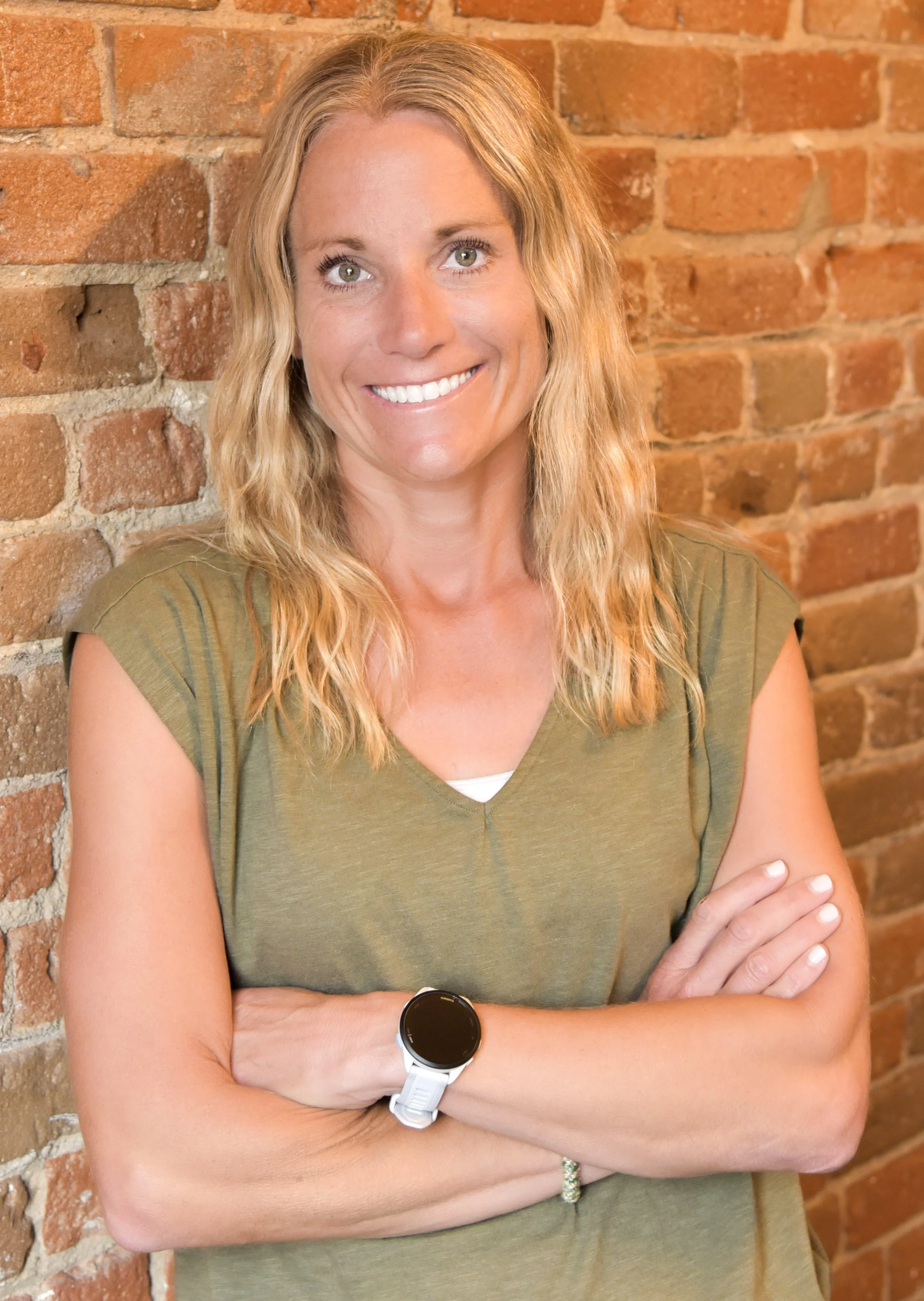 A smiling woman with blonde wavy hair, wearing a green shirt and a white watch, standing with arms crossed against a brick wall background.