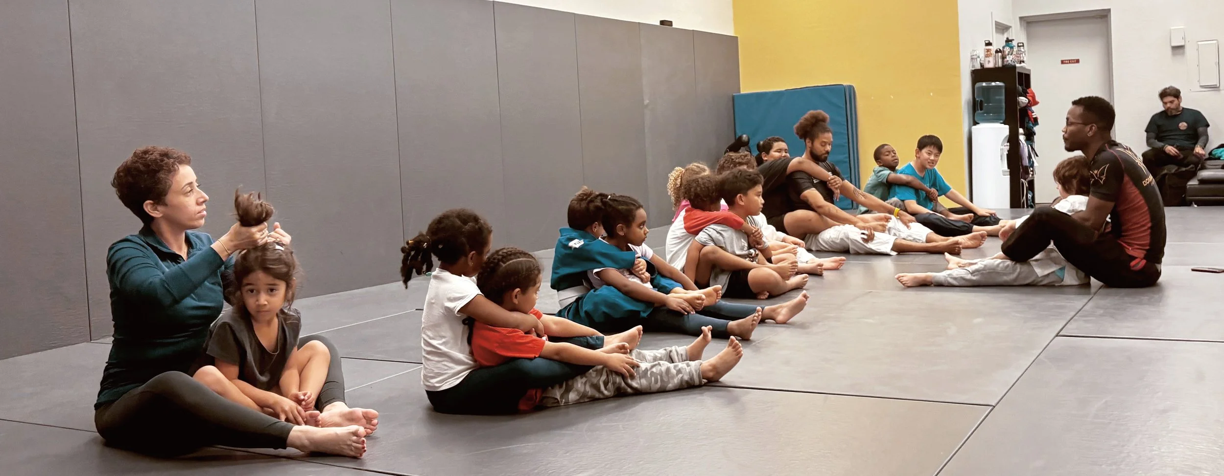 Children and adults sitting on a martial arts training mat, listening to an instructor. The instructor, seated facing the group, is wearing a black shirt with red accents, and two children are sitting against the instructor. The room has gray and yellow walls, and a water cooler is visible in the background.