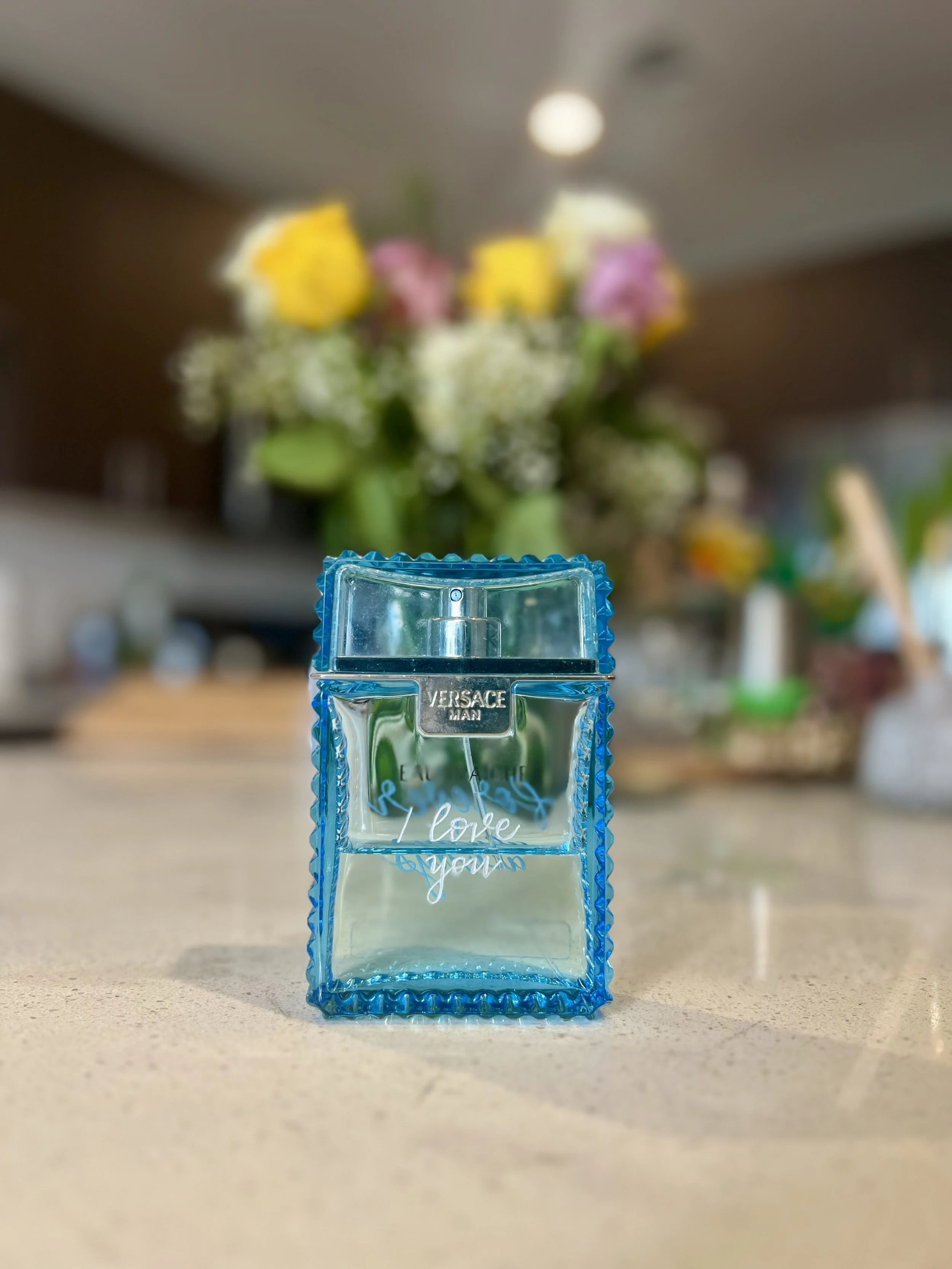 A blue Versace perfume bottle with the words 'I love you' written on it, placed on a beige countertop. In the blurred background, there is a bouquet of multicolored flowers.