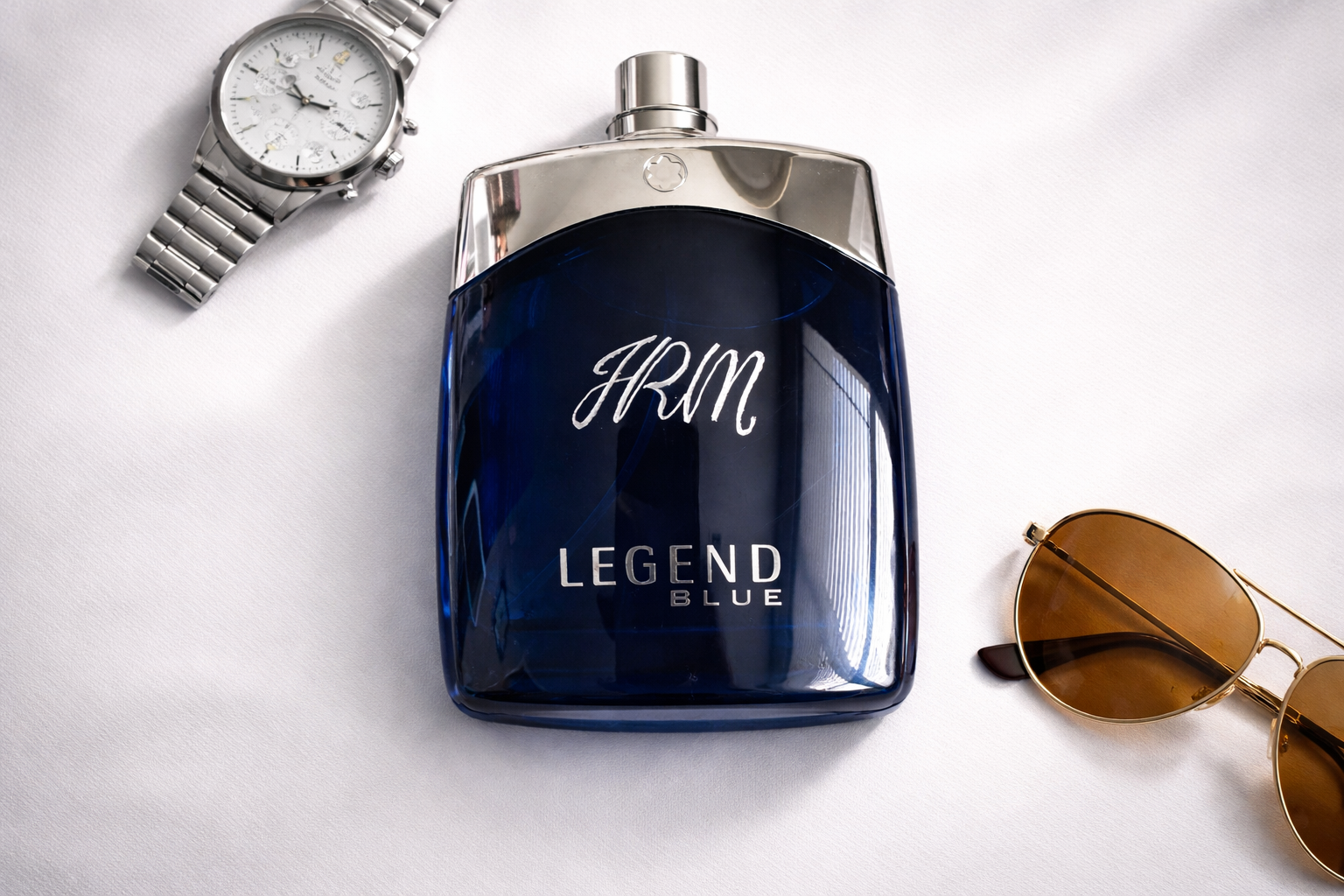 A bottle of Legend Blue by HRM cologne, a silver wristwatch with multiple dials, and gold sunglasses with brown lenses on a white surface.